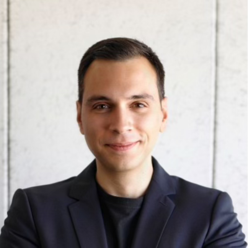 A man with dark hair, wearing a black shirt and a dark blazer, smiling in front of a plain wall.