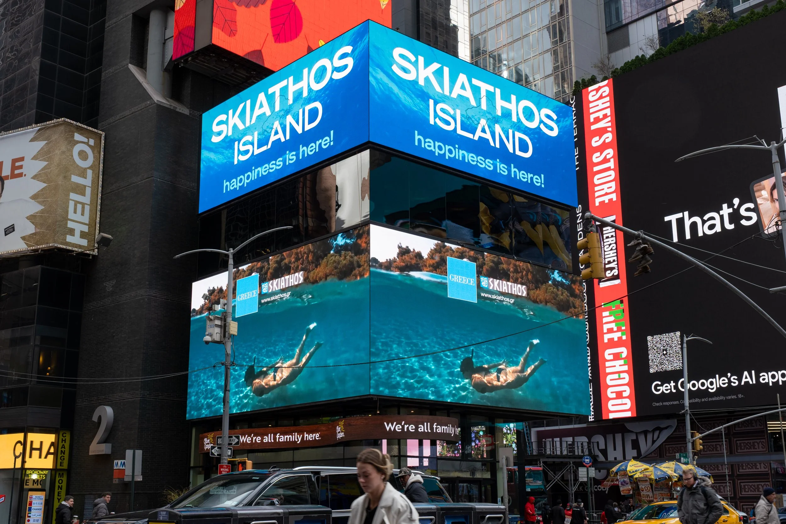 Billboards in Times Square, New York City, featuring advertisements for Skiathos Island in Greece with a large digital screen showing people swimming underwater, and surrounding signs for various brands and stores. Pedestrians and taxis are visible on the street.