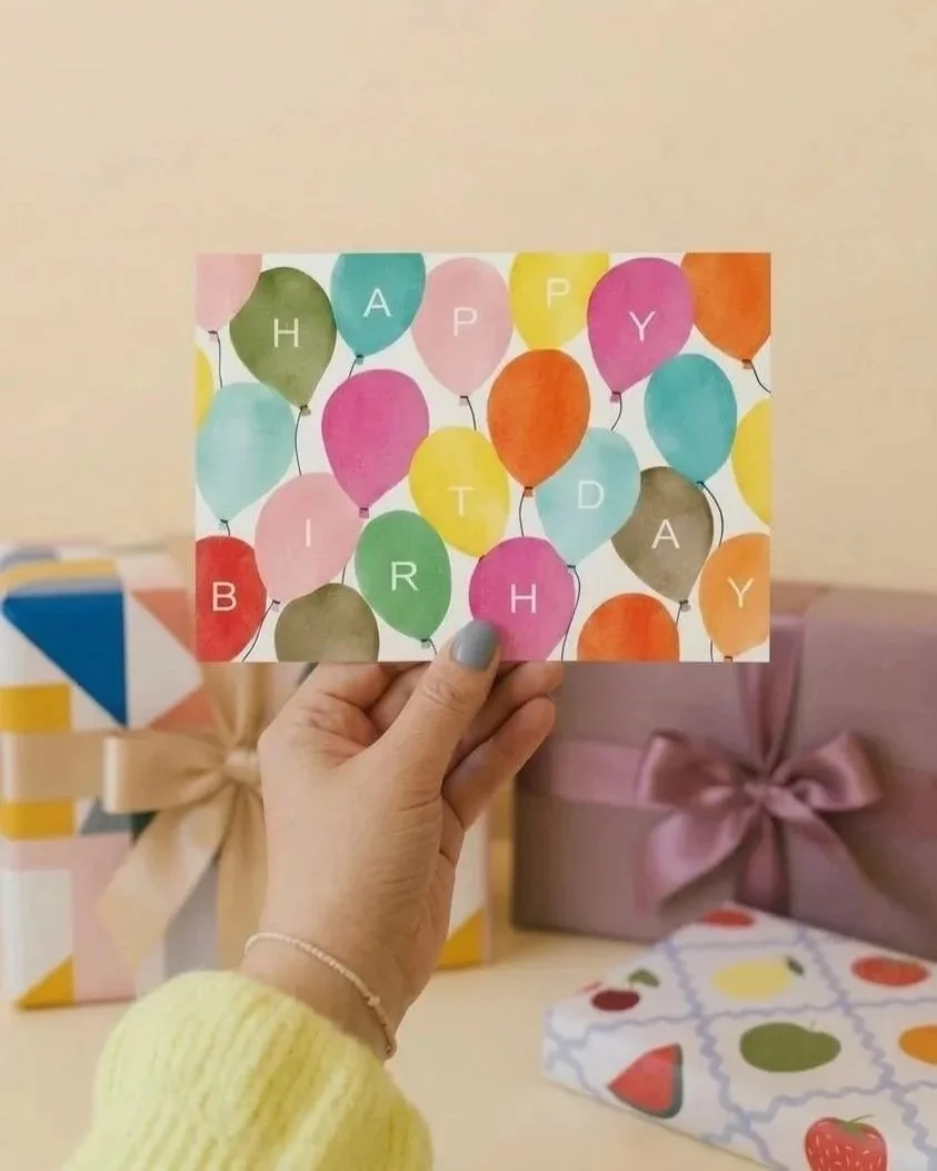 A hand holding a birthday card with colorful balloons spelling out 'HAPPY BIRTHDAY'. In the background, there are wrapped gifts and a table with a patterned wrap.