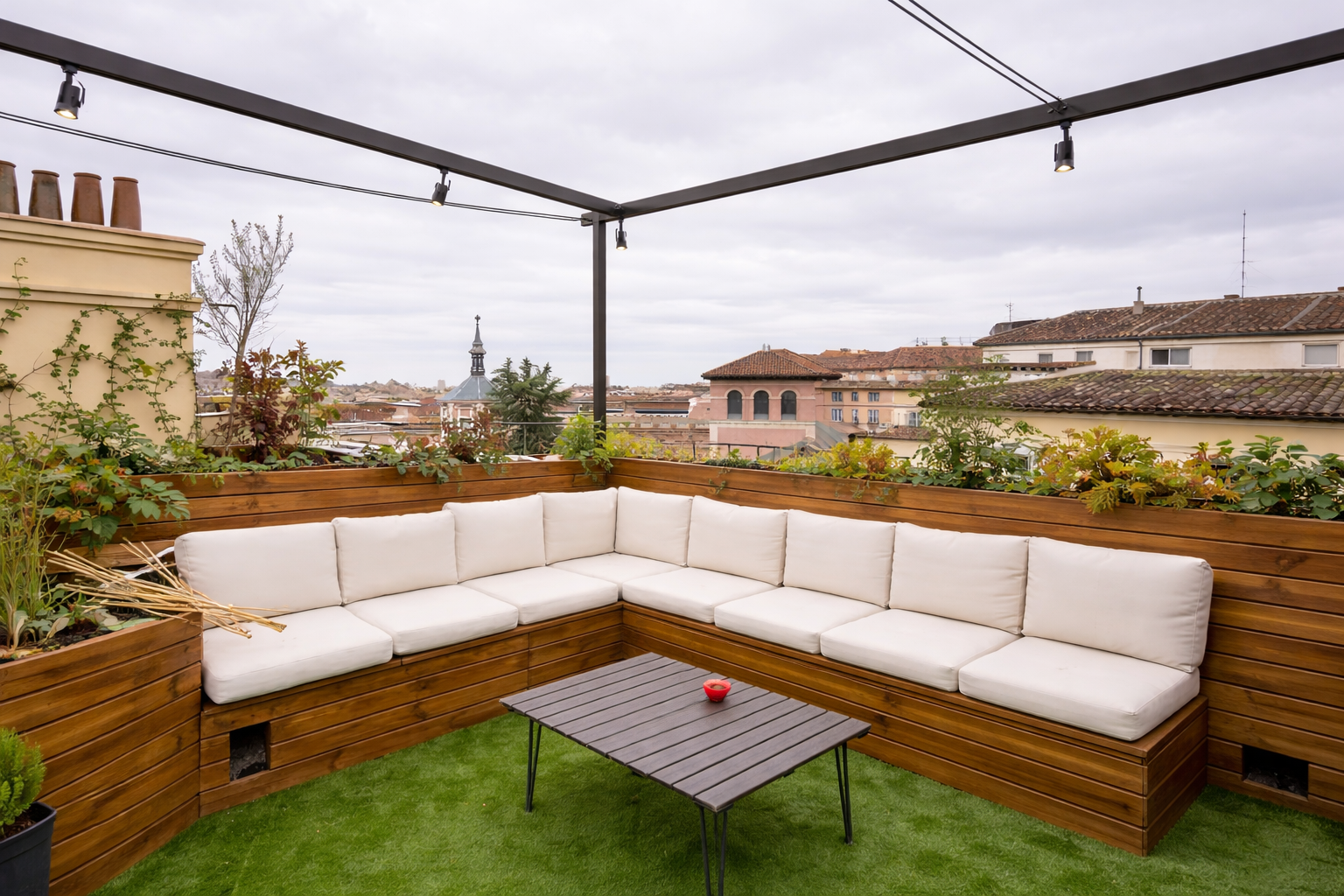 Terraza con esquina de sofá con cojines blancos, mesa pequeña y plantas en un cercado de madera, con vista a techos y edificaciones en un día nublado.