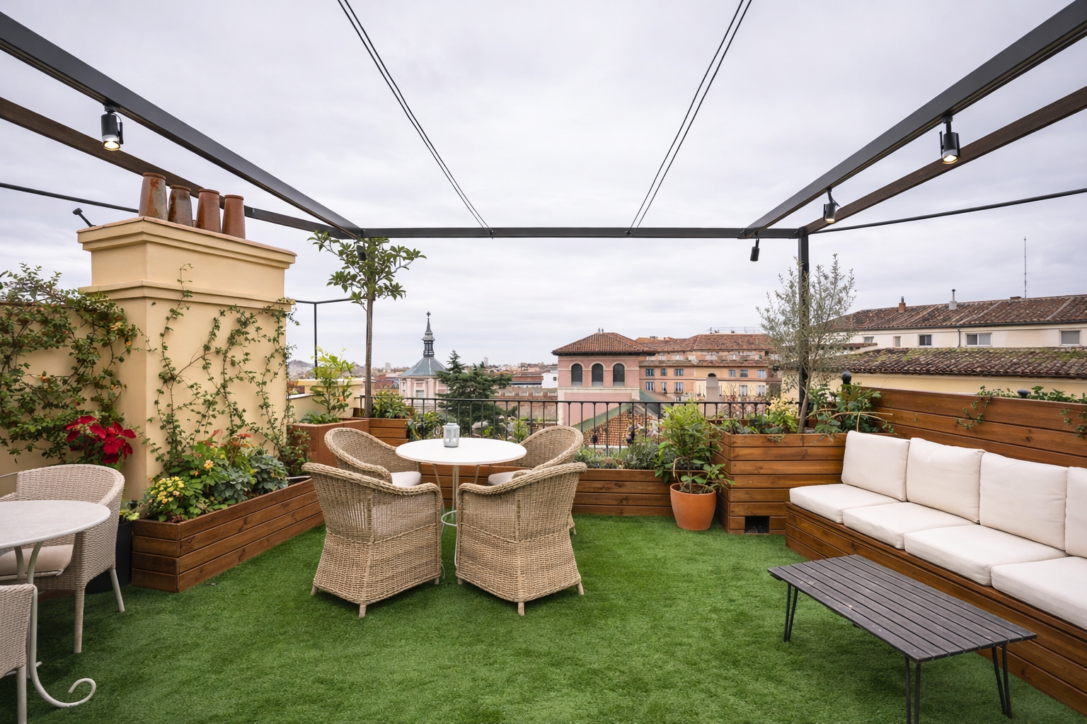 Terraza en la azotea con muebles de mimbre y cojines blancos, plantas en jardineras de madera y piso de césped artificial, con vista urbana de edificios y una iglesia en el fondo, cielo nublado.