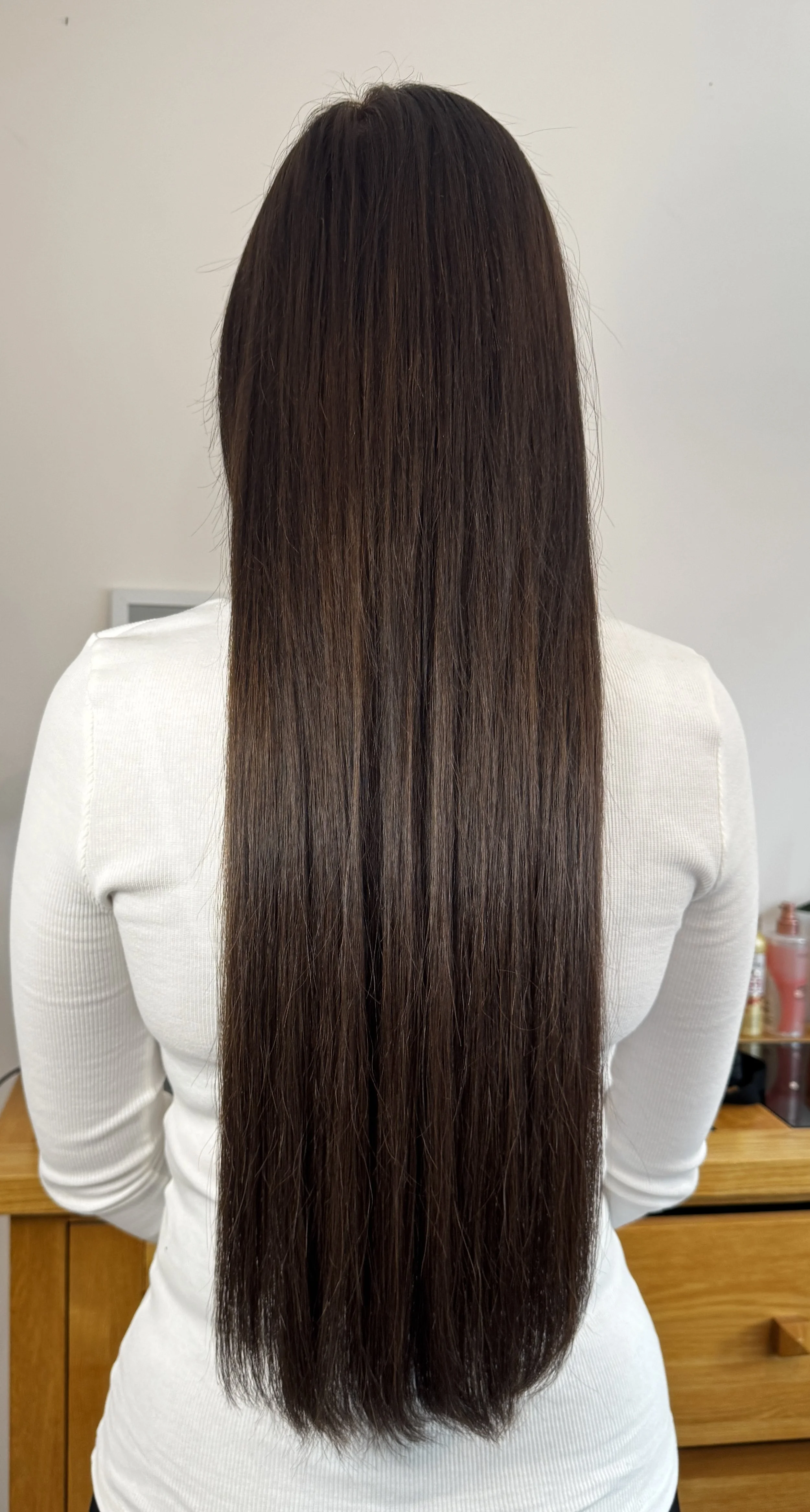 Back view of a person with long, straight brown hair, wearing a white long-sleeve shirt, standing in a room with a wooden dresser and a few bottles on top.
