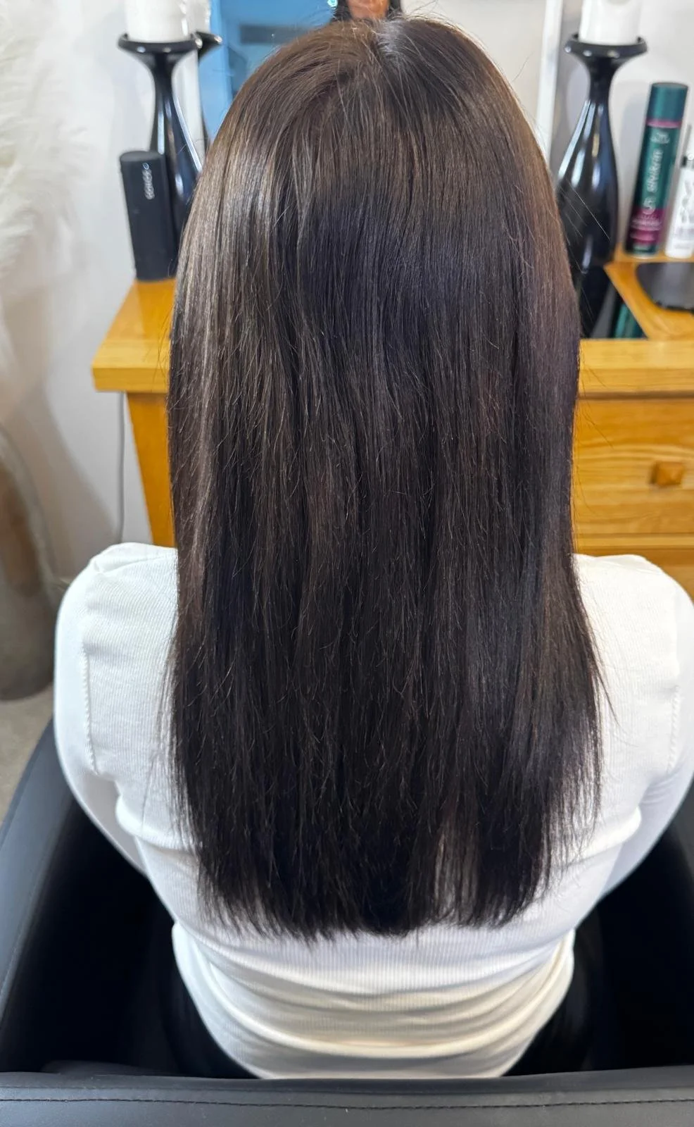 Back view of a woman with long, straight, dark brown hair sitting in a salon chair.