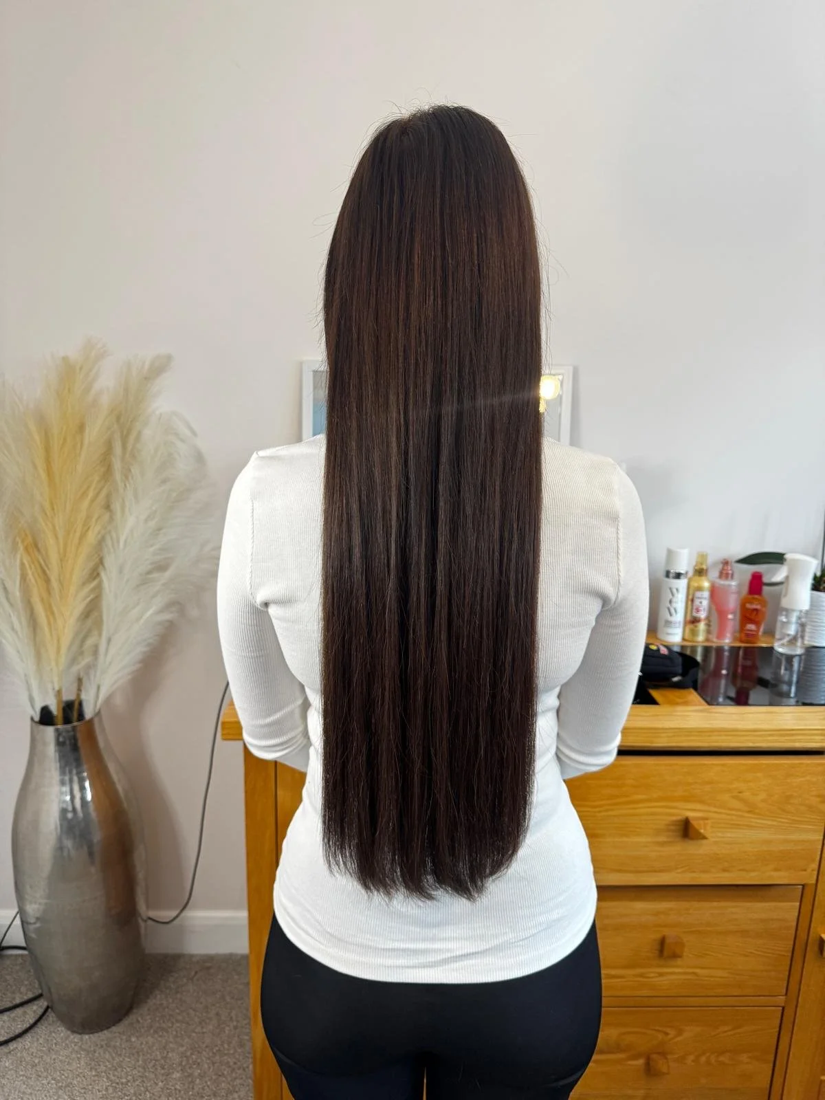 Back view of a woman with long, straight, dark brown hair, standing in a room with a white wall, a wooden dresser, and a decorative vase with pampas grass.