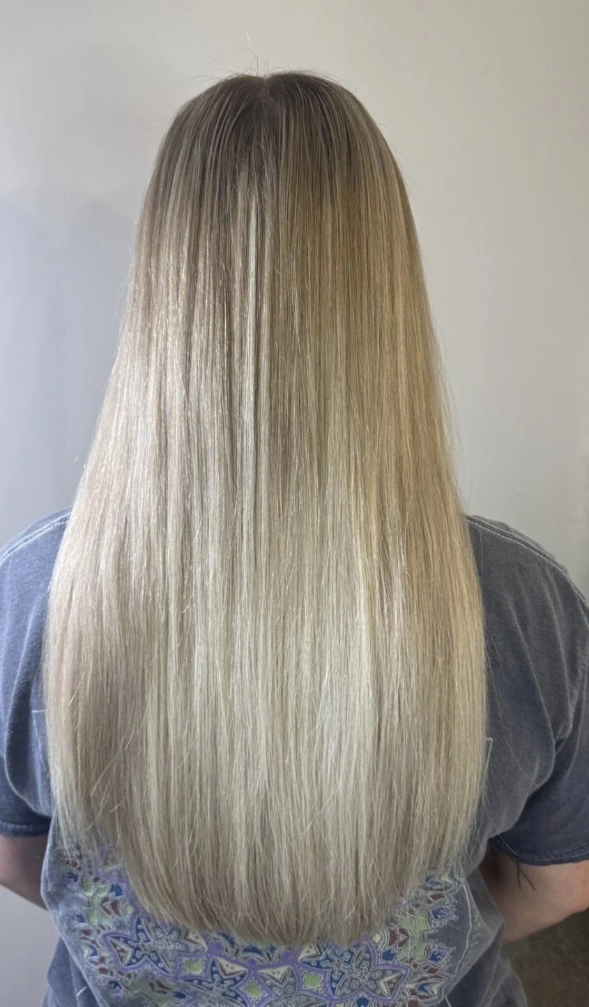 Back view of a person with long, straight blonde hair, wearing a grey shirt with colorful patterns.