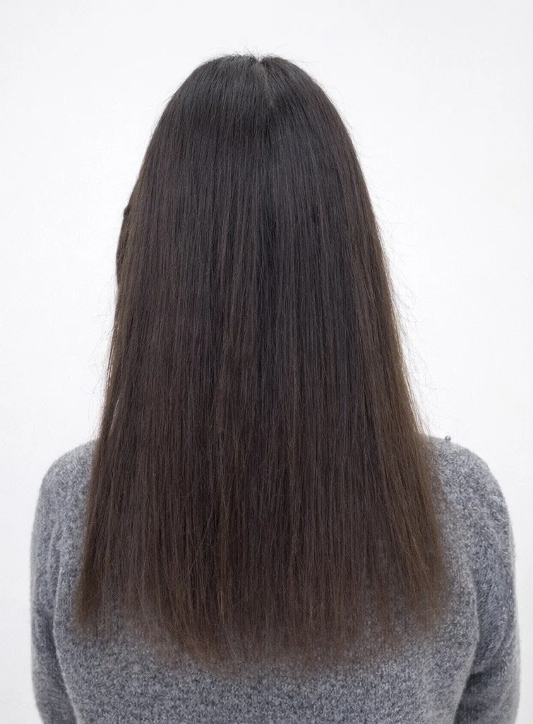 Back view of a person with long, straight brown hair wearing a gray sweater against a plain white background.