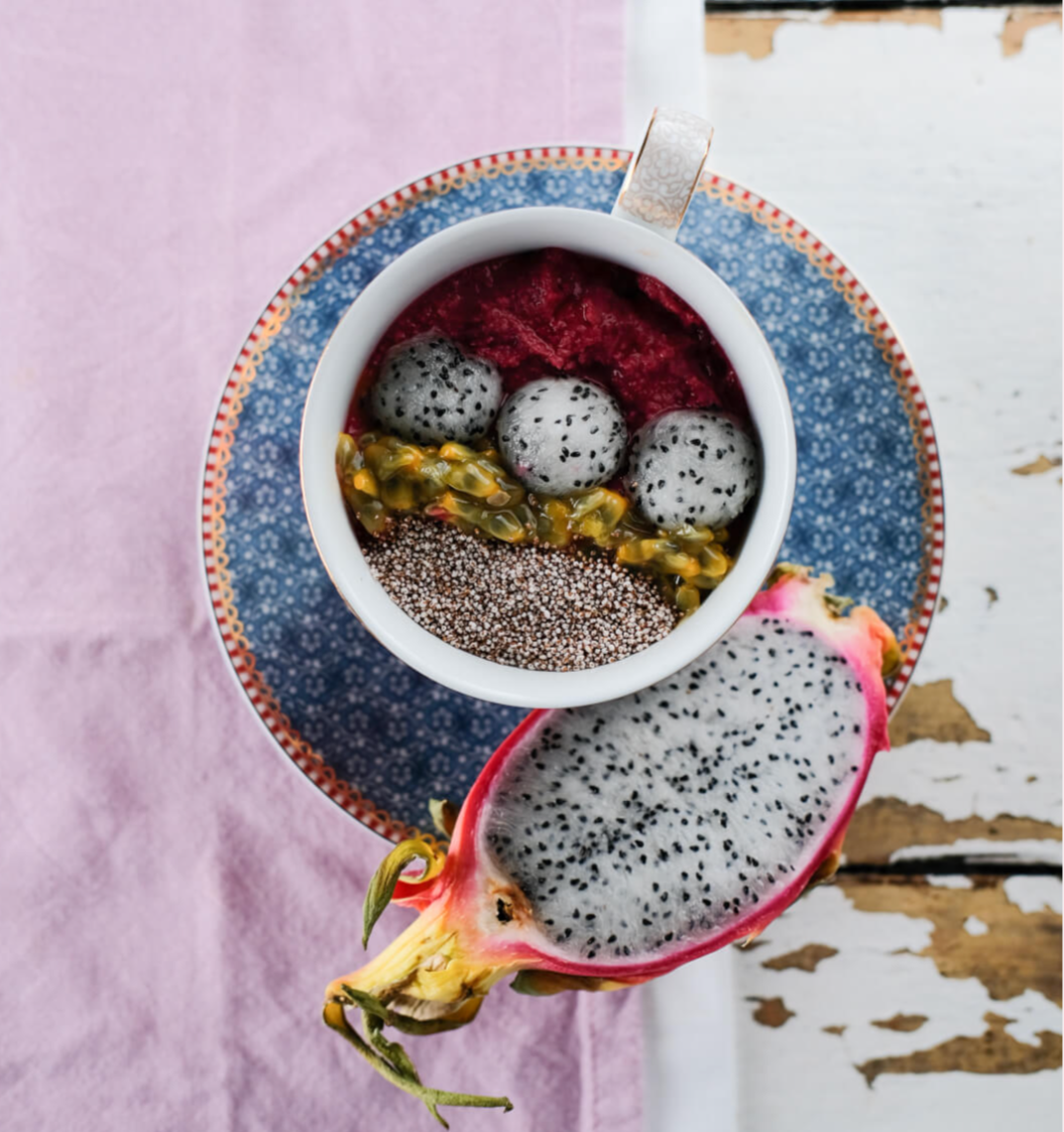 A cup of smoothie or yogurt topped with dragon fruit balls, passion fruit, seeds, and a half dragon fruit with white flesh and black seeds on a decorative plate, with a pink cloth and worn white wooden surface.