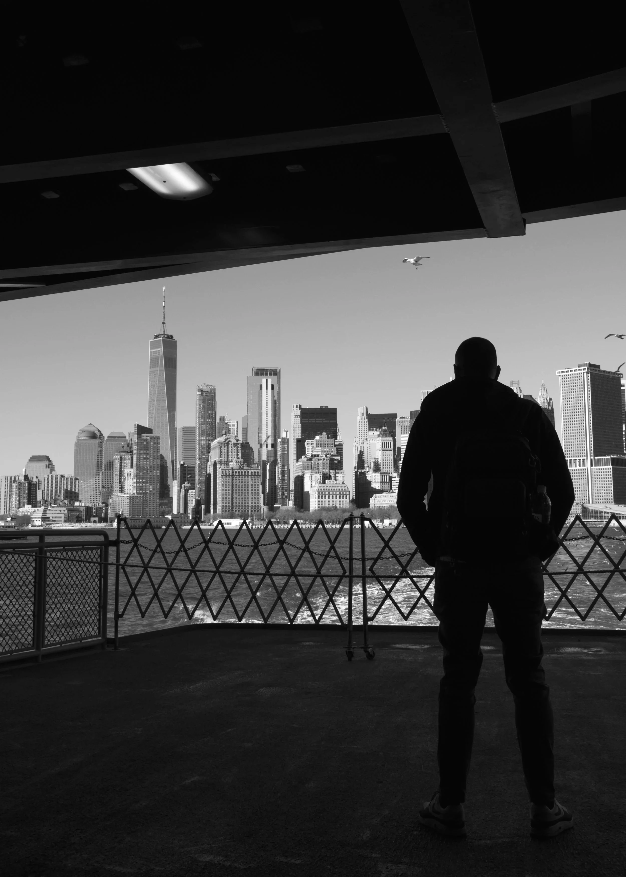 Escape. Black and white photograph of a figure standing on the stern of a ferry with a city skyline in the distance.