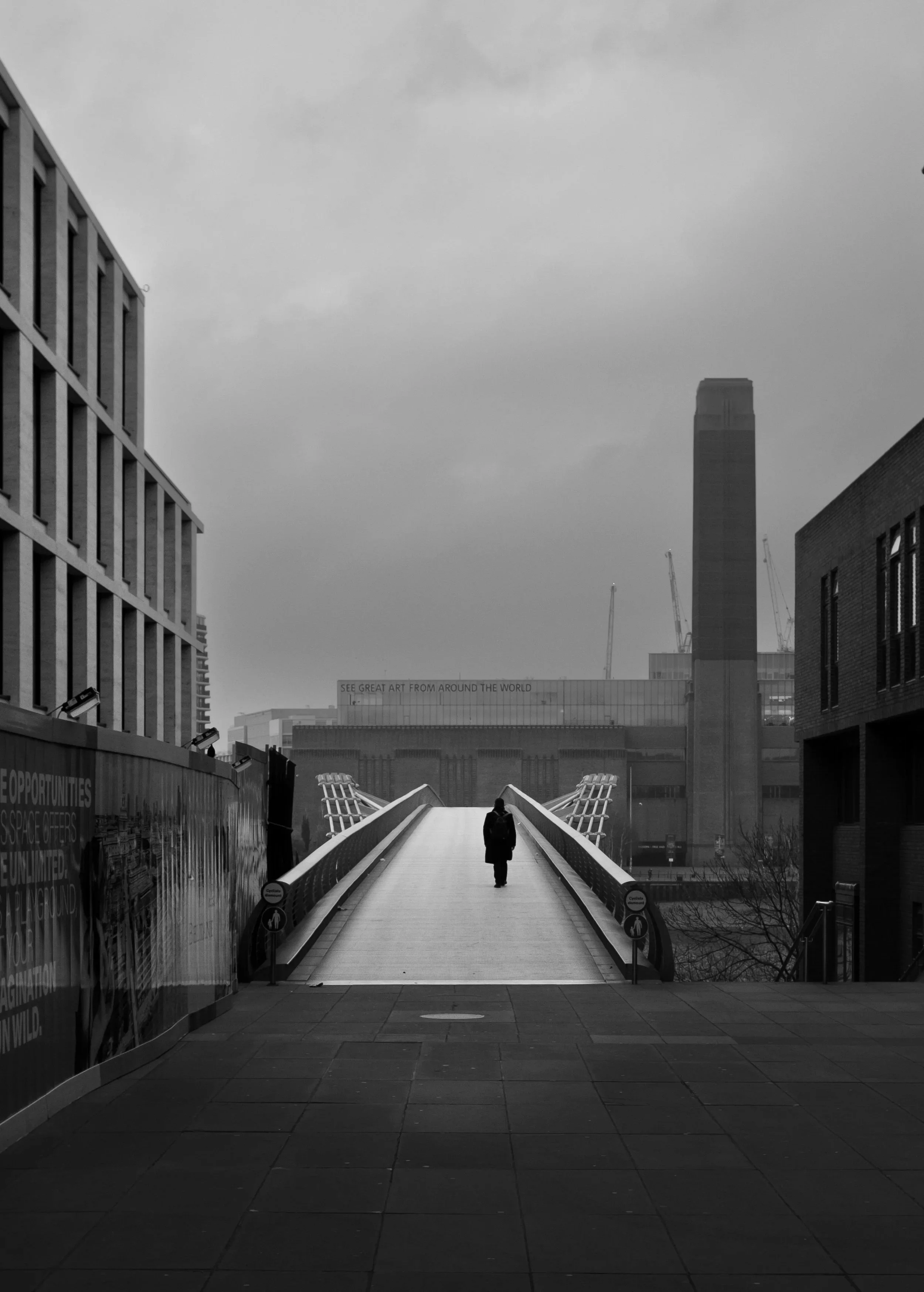 Over. Black and white photograph of a solitary figure walking across a bridge spanning a river.