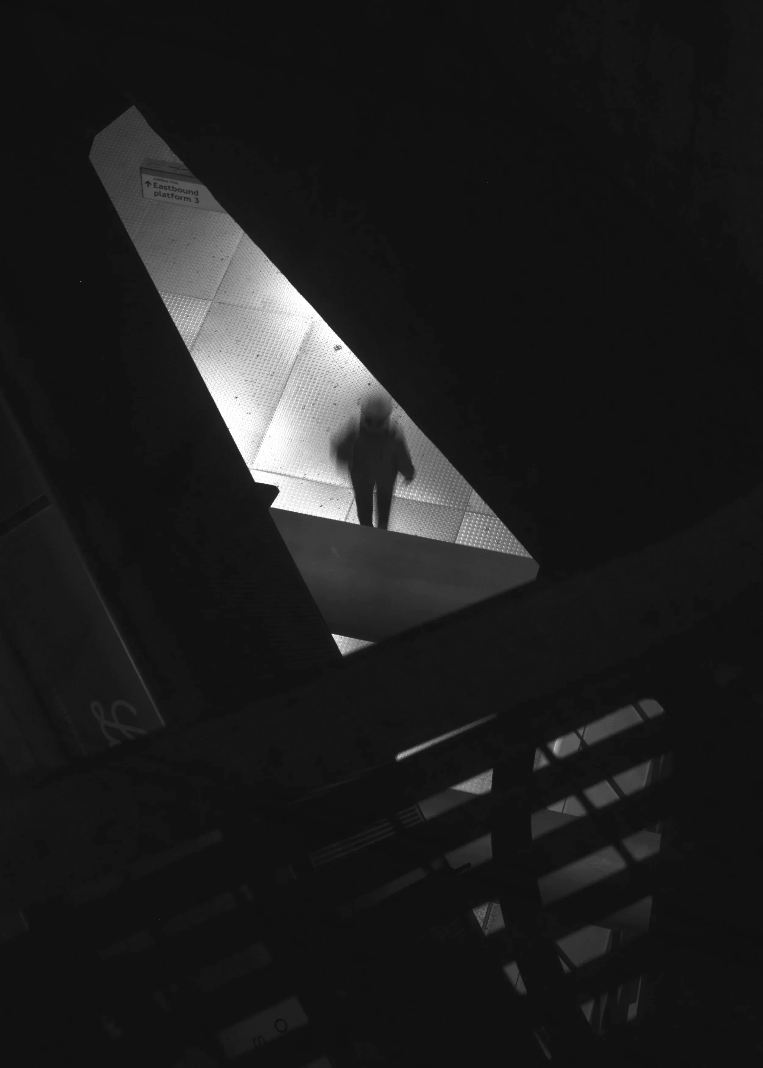 Eastbound. Black and white photograph of a blurred figure in an underground station beneath diagonal light and signage. 