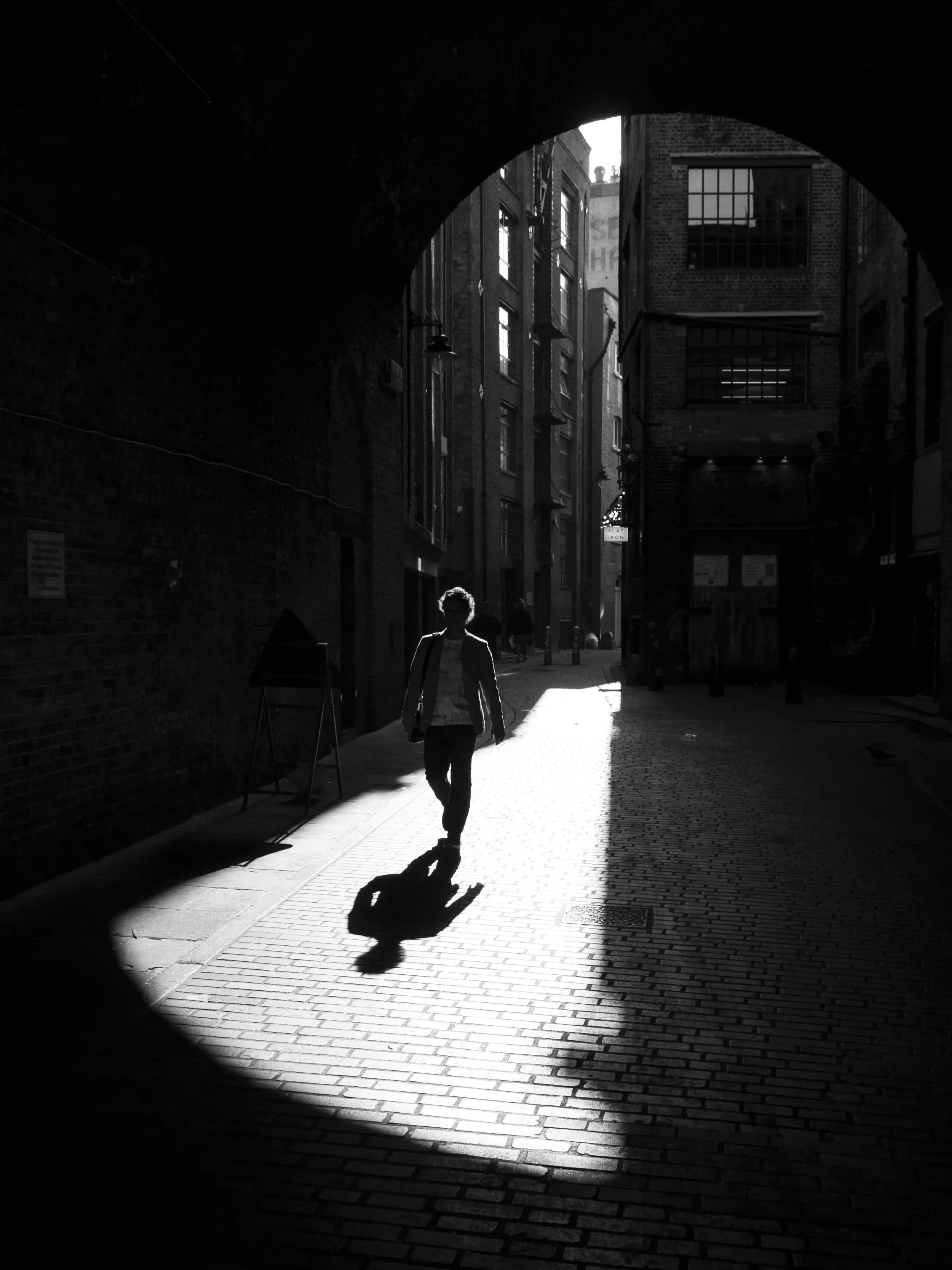 Enlightened. Black and white photograph of a figure walking out of a dark alley illuminated by a single shaft of light.