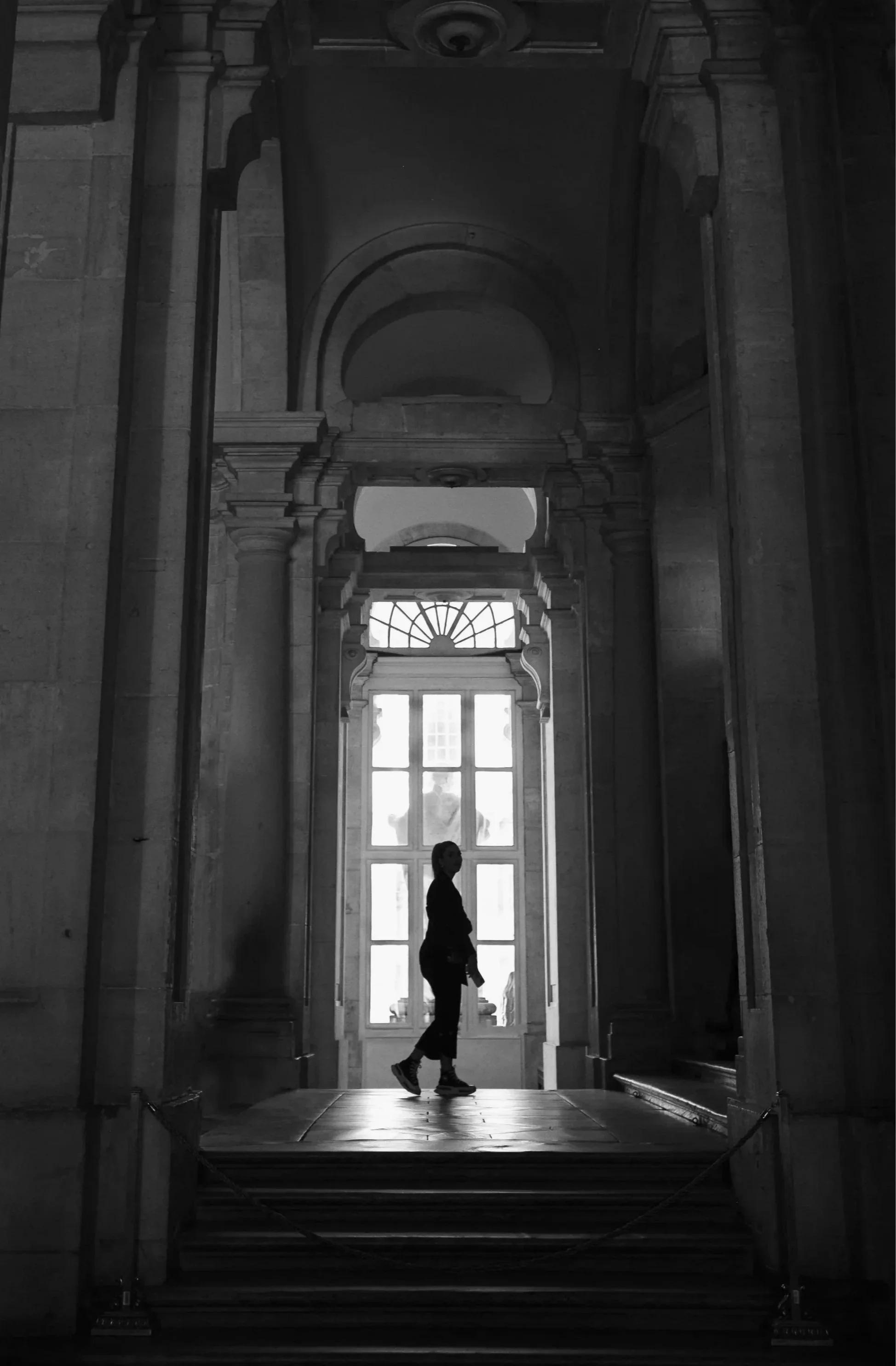 Grand Entrance. Black and white photograph of a solitary figure at the entrance of a grand collonaded corridor with tall arched windows.