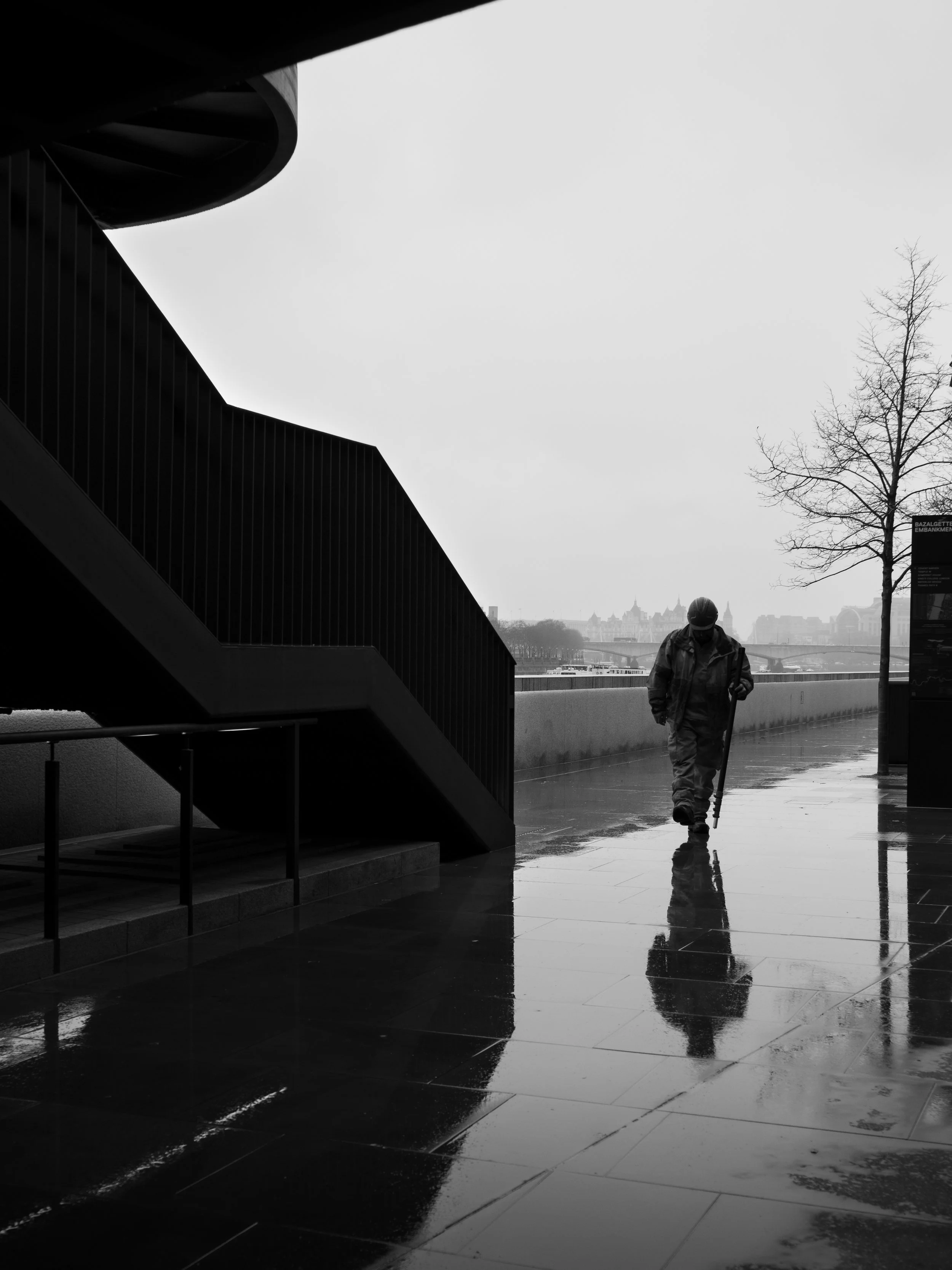A person walking with a cane on a wet sidewalk beneath modern architecture, with a tree and cityscape visible in the distance on a rainy day in black and white.