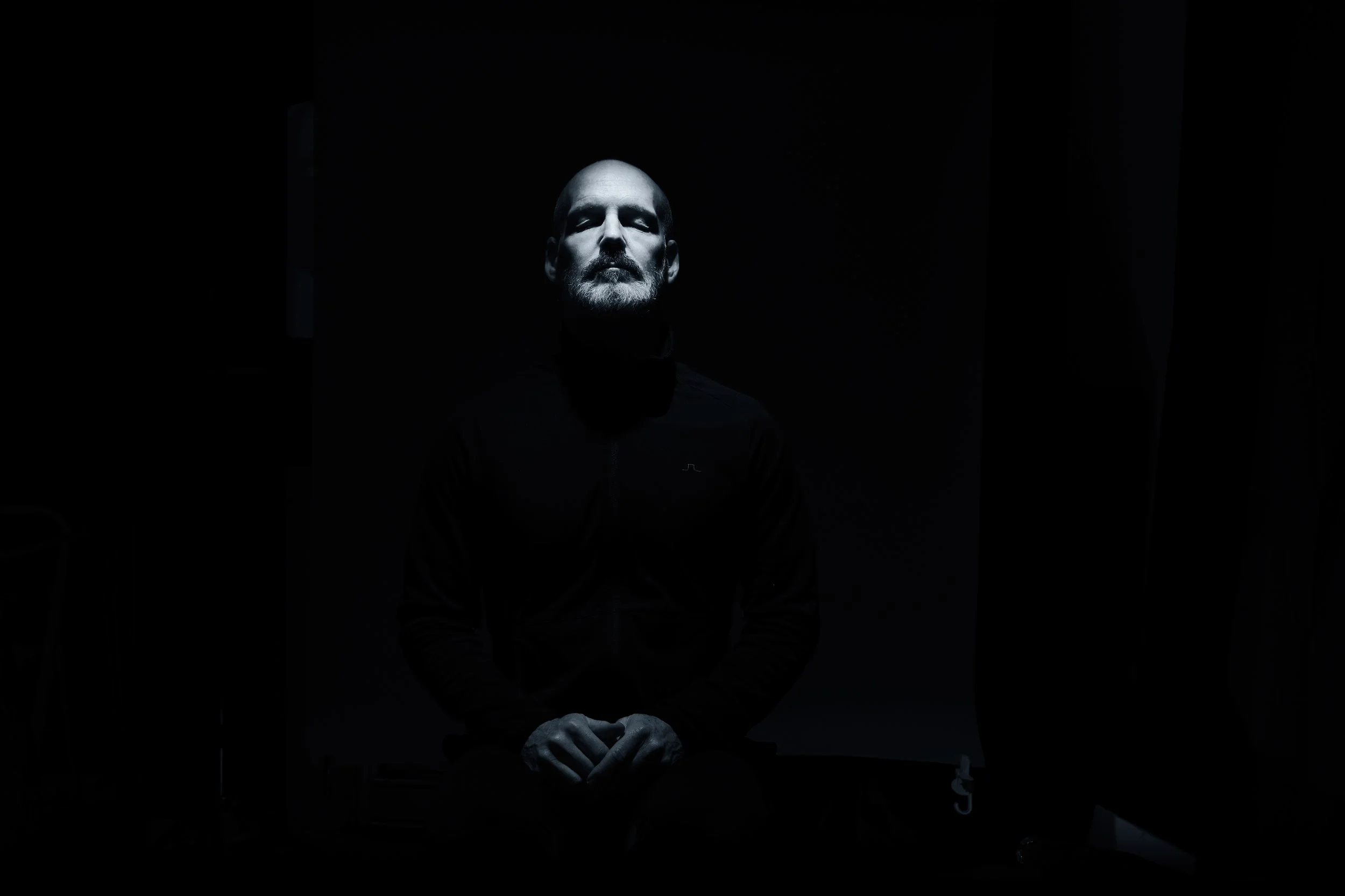 A man with a shaved head and beard sitting in darkness, illuminated from above, wearing a black jacket.