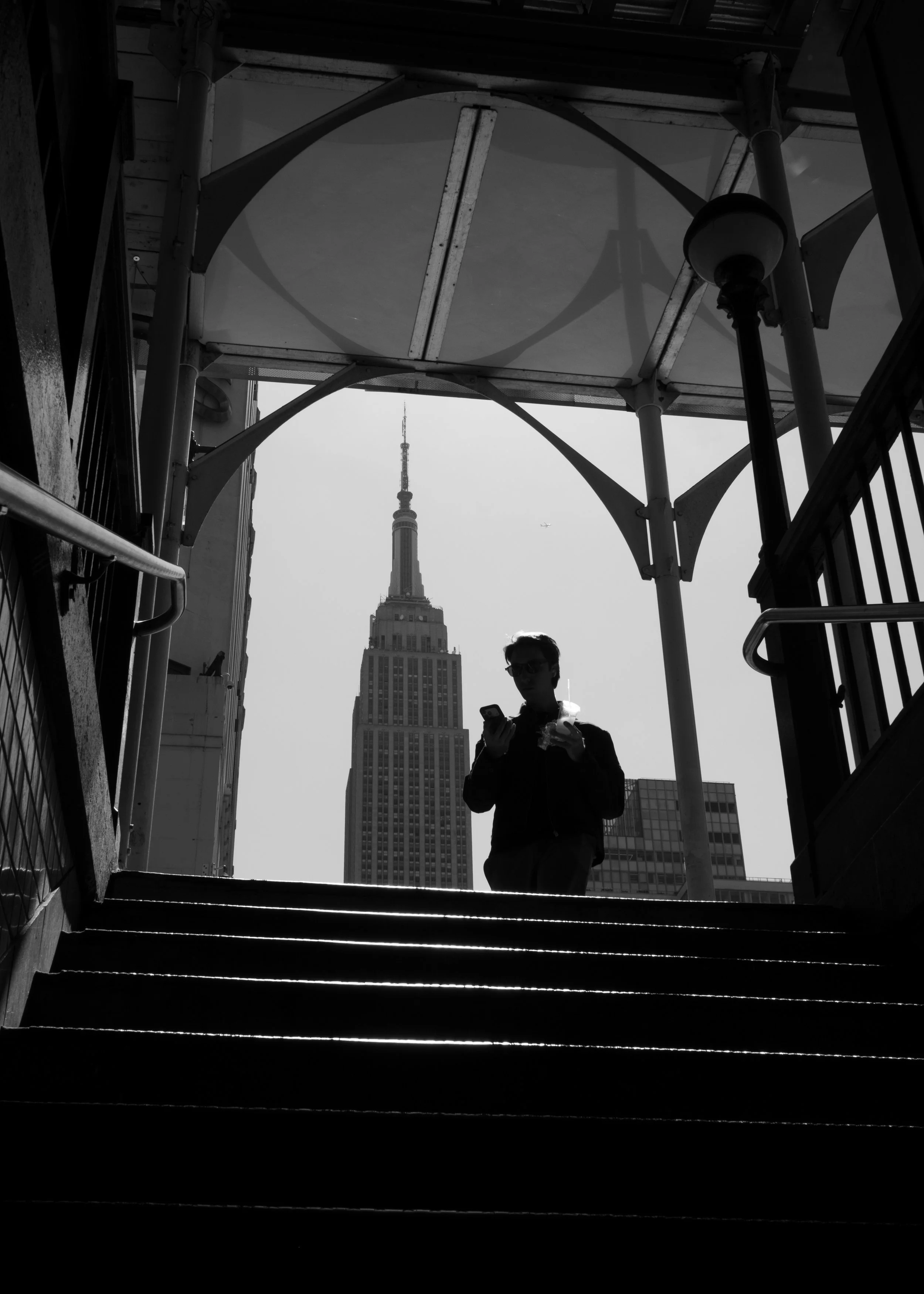 Empire Rising. Black and white photograph of a silhouetted figure approaching a staircase with the Empire State Building visible through an iron framework above.