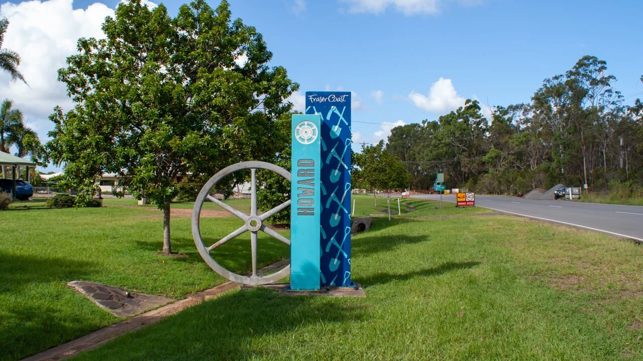 Sign for Hervey and Fraser Coast regions with a decorative wheel and blue artwork on a grassy roadside