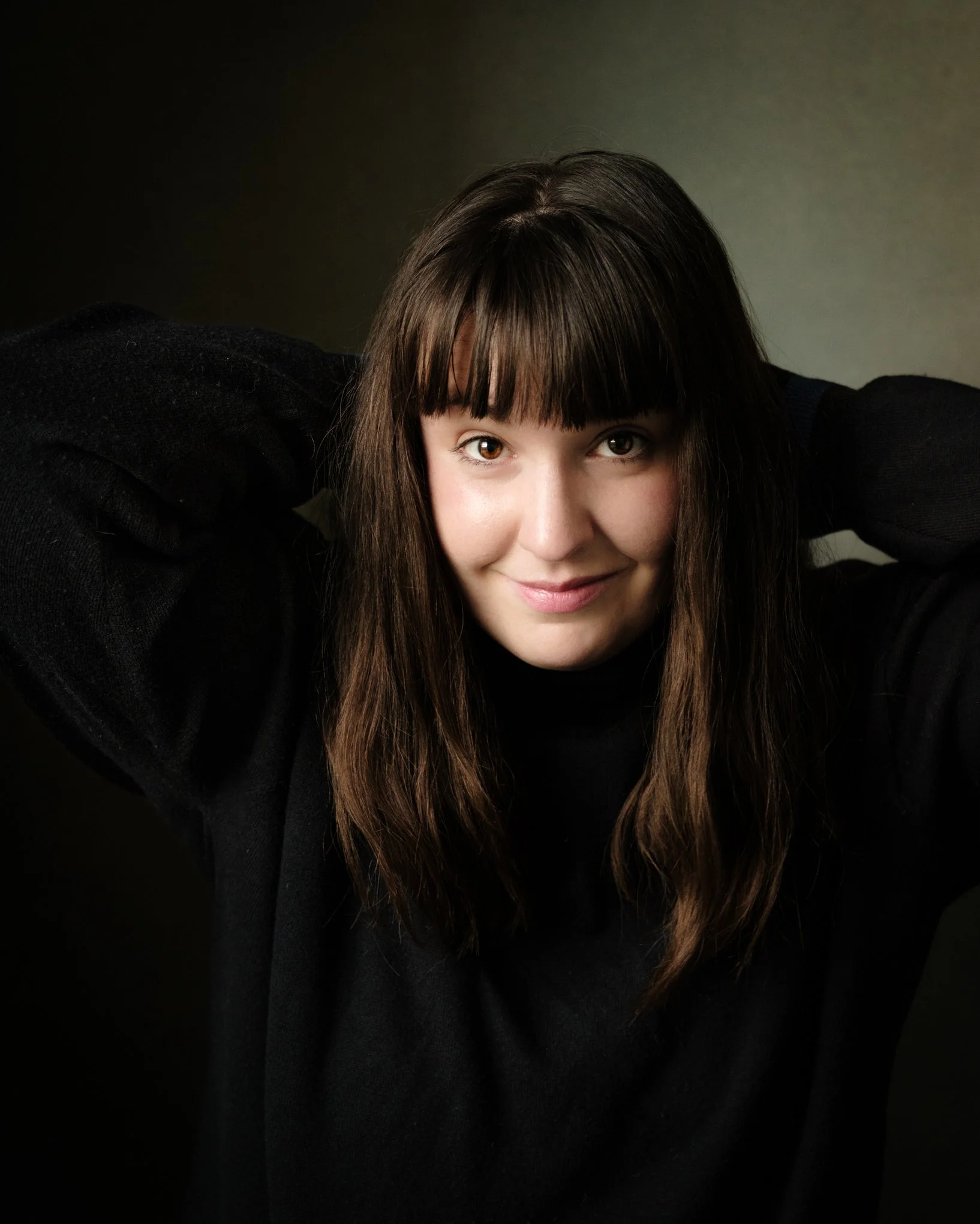 A woman with long brown hair and bangs, smiling slightly, with her hands behind her head, wearing a black top, against a dark background.