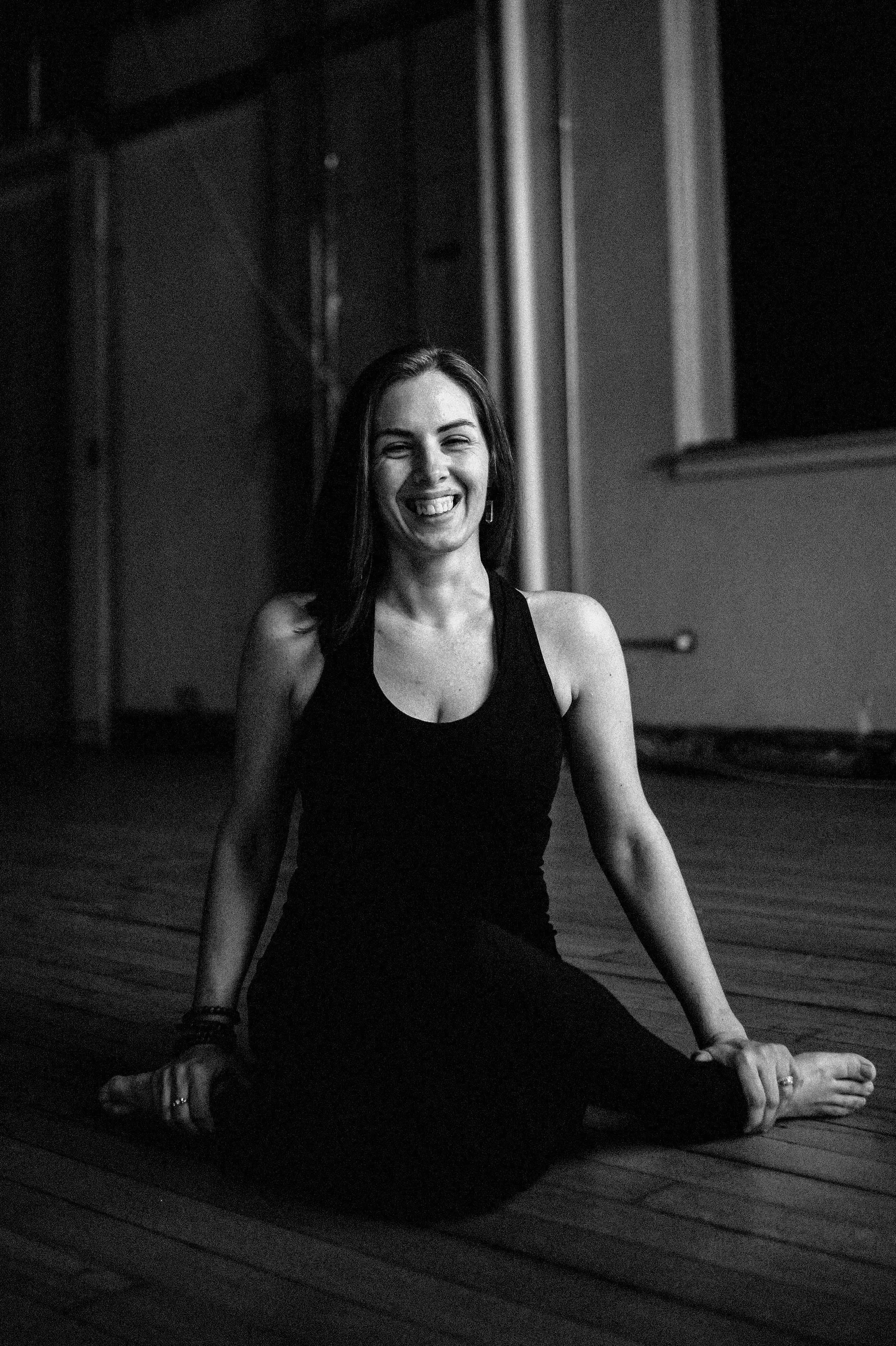 A smiling woman with dark hair sitting on a wooden floor in a black sleeveless top and black pants, in a dimly lit room.