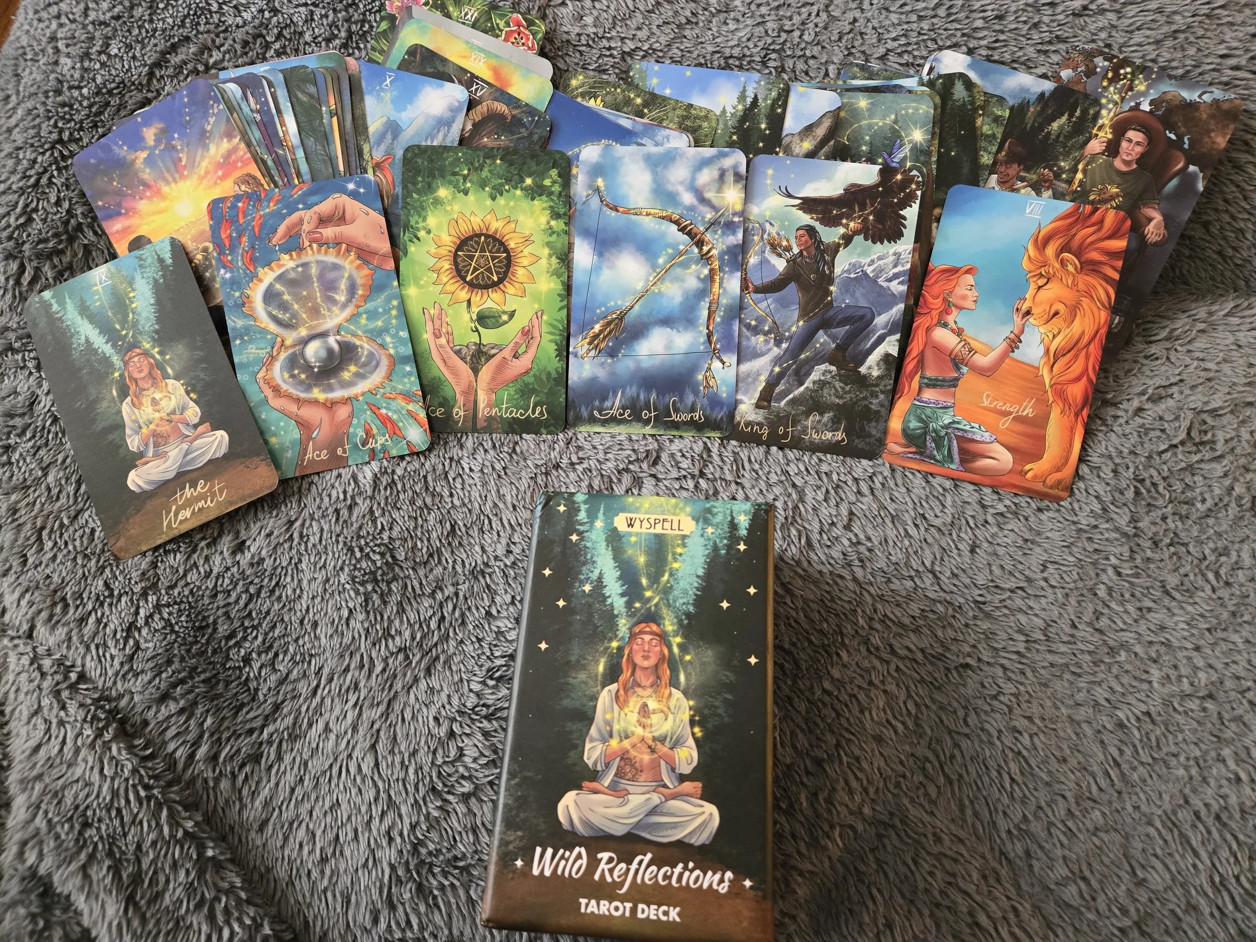 A collection of colorful tarot cards and a tarot deck box called "Wild Reflections" arranged on a gray furry surface. The cards depict mystical and fantasy-themed imagery, including a woman meditating, magical symbols, a sunflower, a bow and arrow, a woman with a lion, and a woman with a ladybug. The cards include titles like "the Hermit," "Ace of Cups," "Ace of Pentacles," "Ace of Swords," "King of Swords," and "Strength."