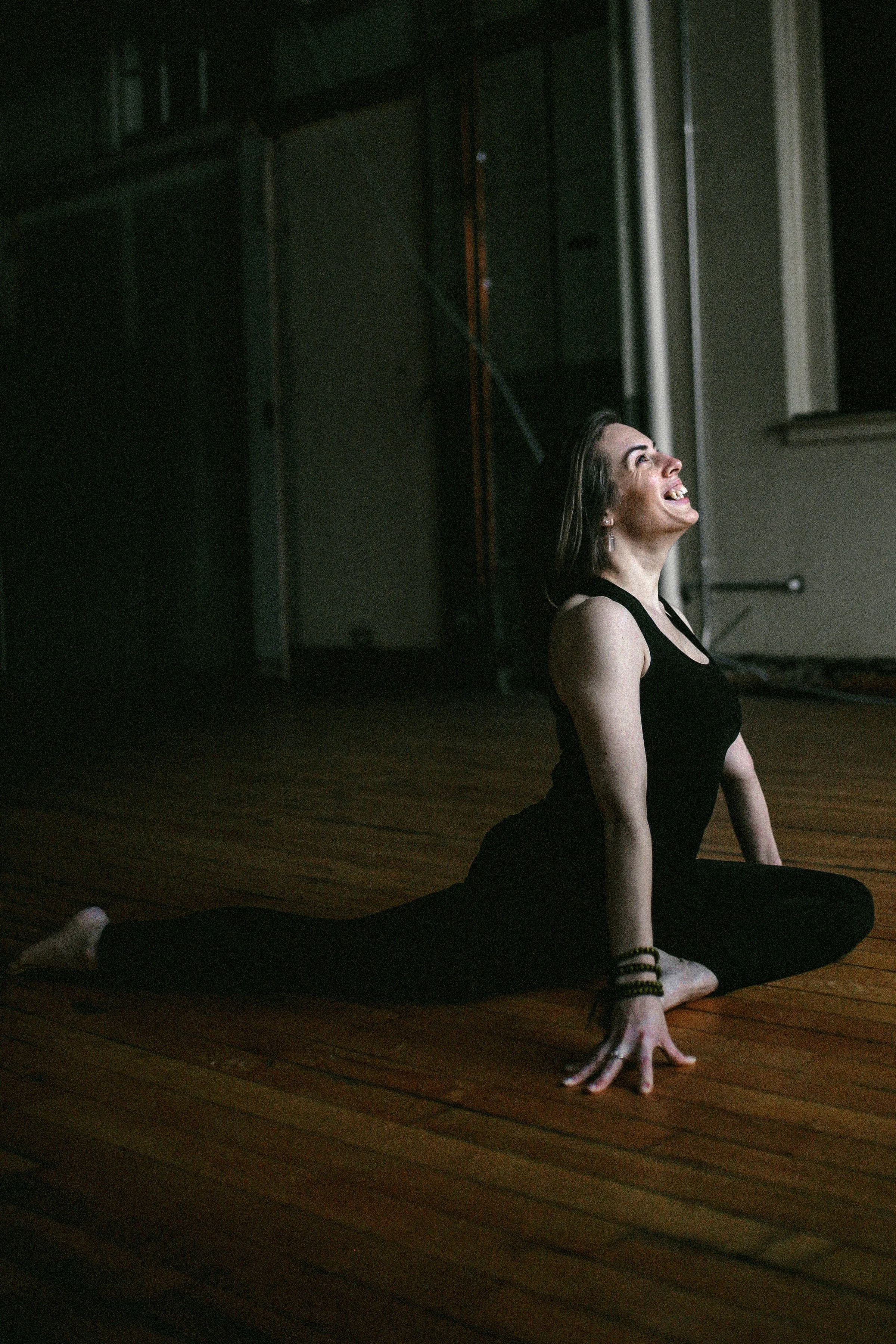 A woman practicing yoga on a wooden floor, smiling and stretching in a studio with dim lighting.