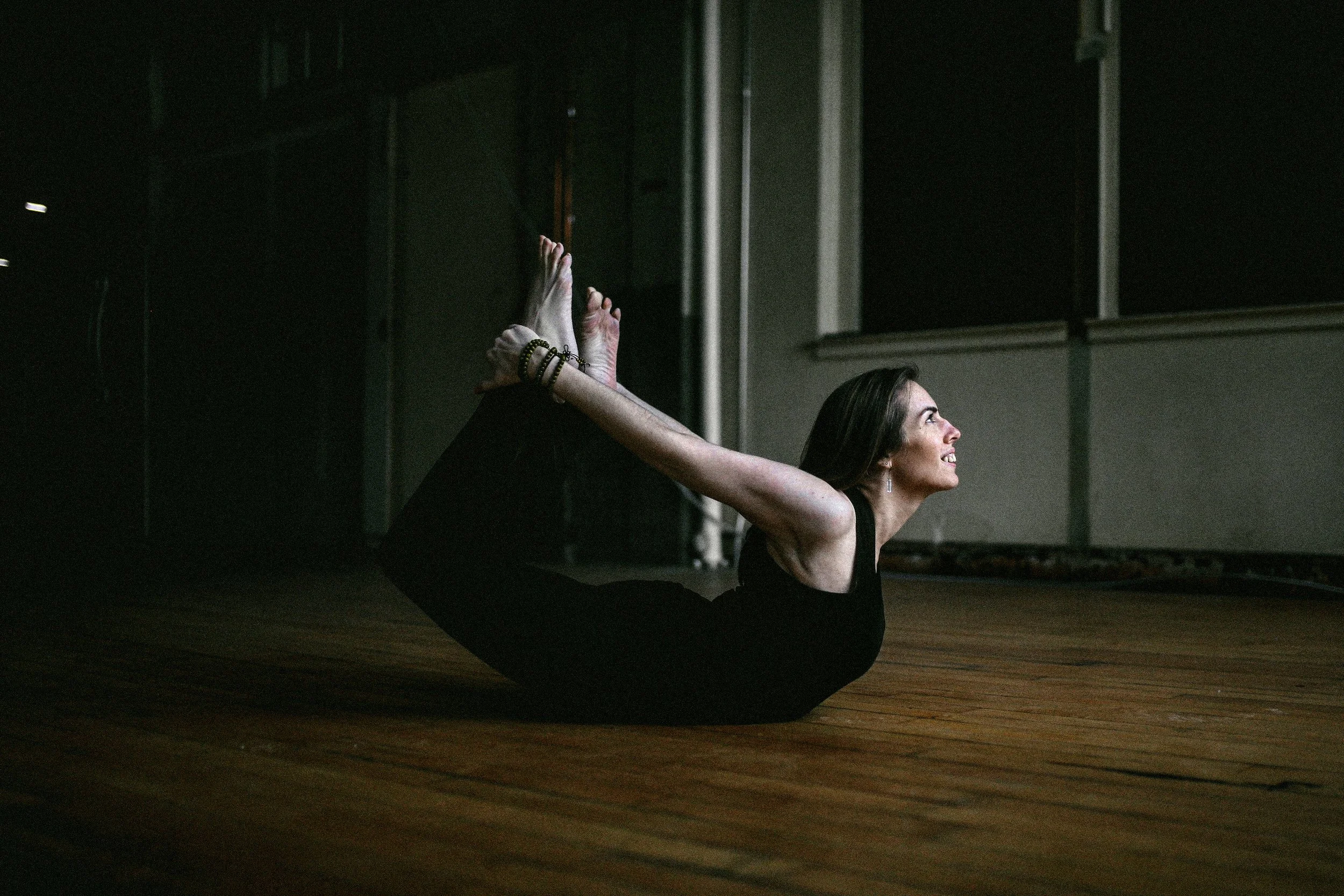 A woman practicing yoga on a wooden floor, lying on her stomach with her arms extended backward, grasping her ankles, and smiling as she looks up.