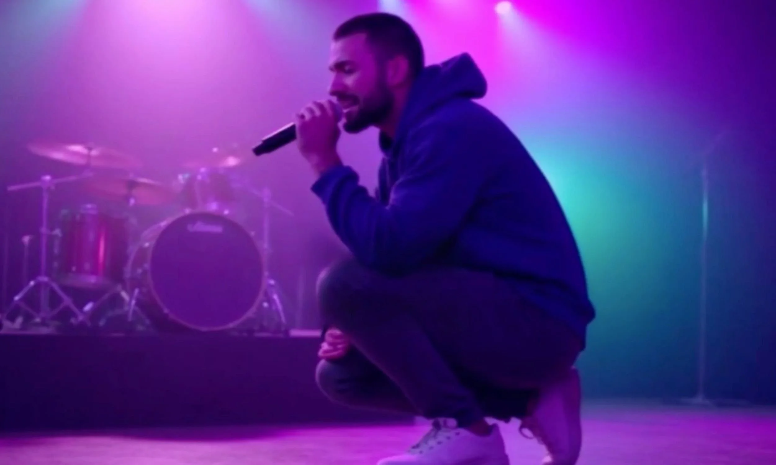 JT Catalano crouched on stage holding a microphone, with a drum set and colorful stage lights in the background performing live.