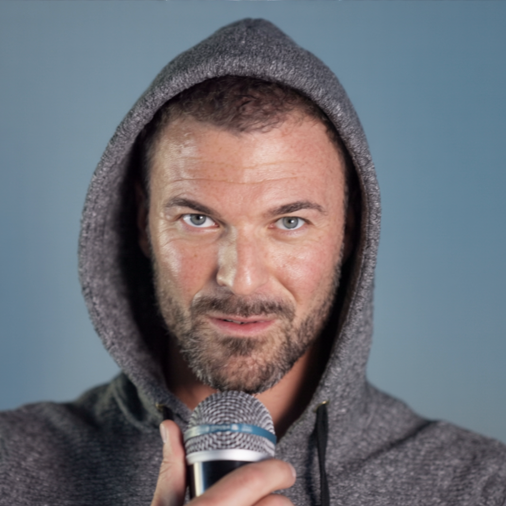 A man with a beard and blue eyes wearing a gray hoodie, holding a microphone close to his mouth, looking directly at the camera.