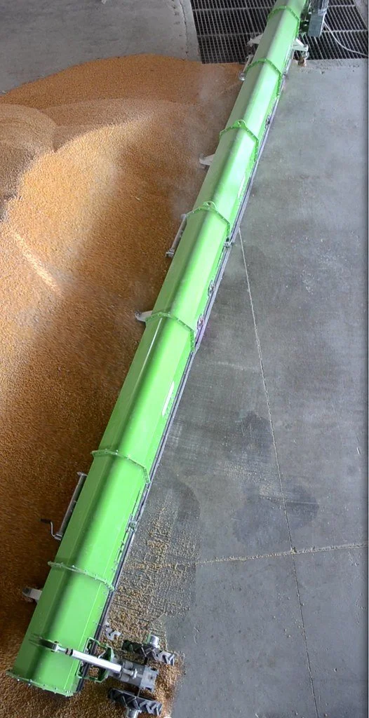 A large green grain conveyor with a moving belt system unloading a pile of grain, likely at a farm or industrial facility.