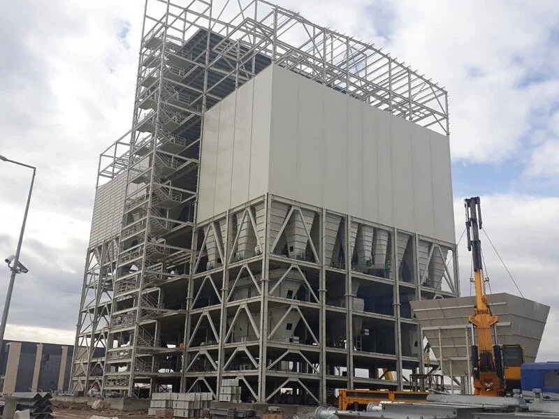 Construction site with a large industrial building supported by a steel framework, including a crane on the right side.