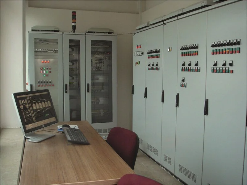 Laboratory control room with electrical panel cabinets, a desk, a computer monitor, and chairs.