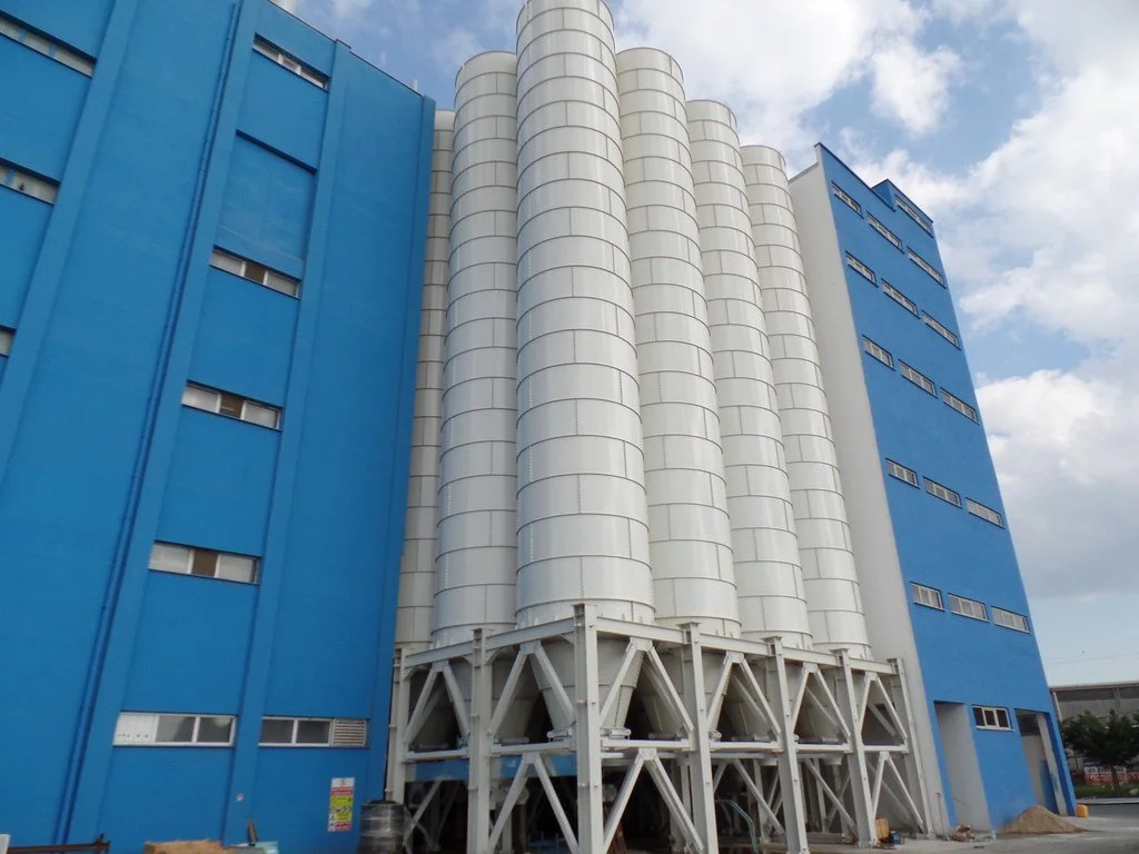 A large blue industrial building with white cylindrical tanks on the outside, set against a partly cloudy sky.