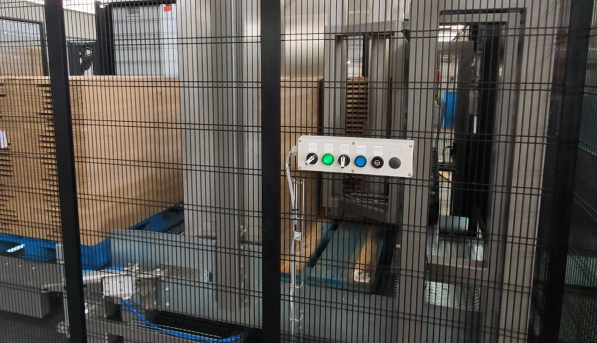 Industrial machine enclosed in a black wire mesh fencing, with control panel featuring green, blue, black, and gray buttons, surrounded by large cardboard boxes on wooden pallets.