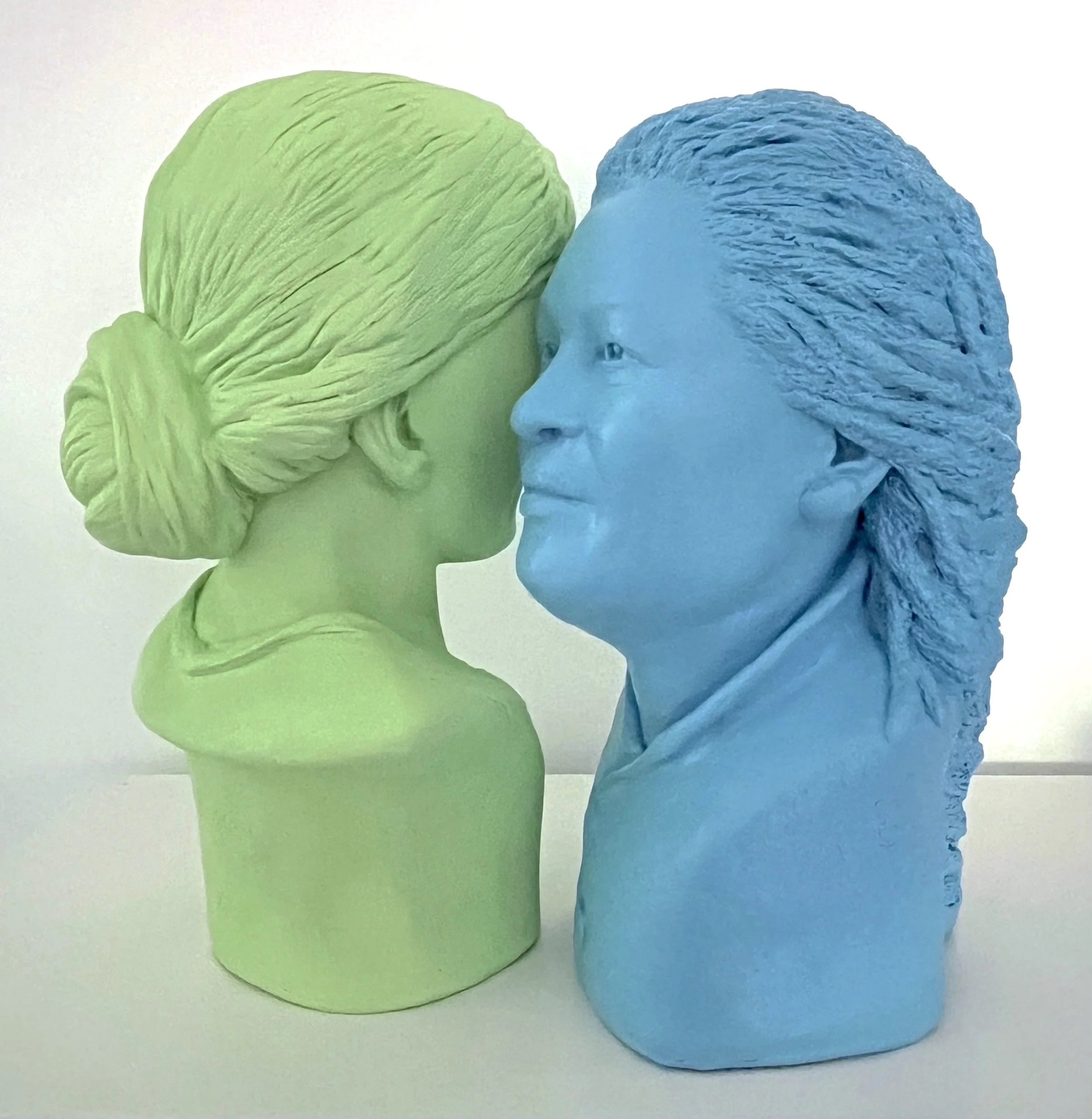 Green and blue bust sculptures of two women facing each other, placed on a white surface.