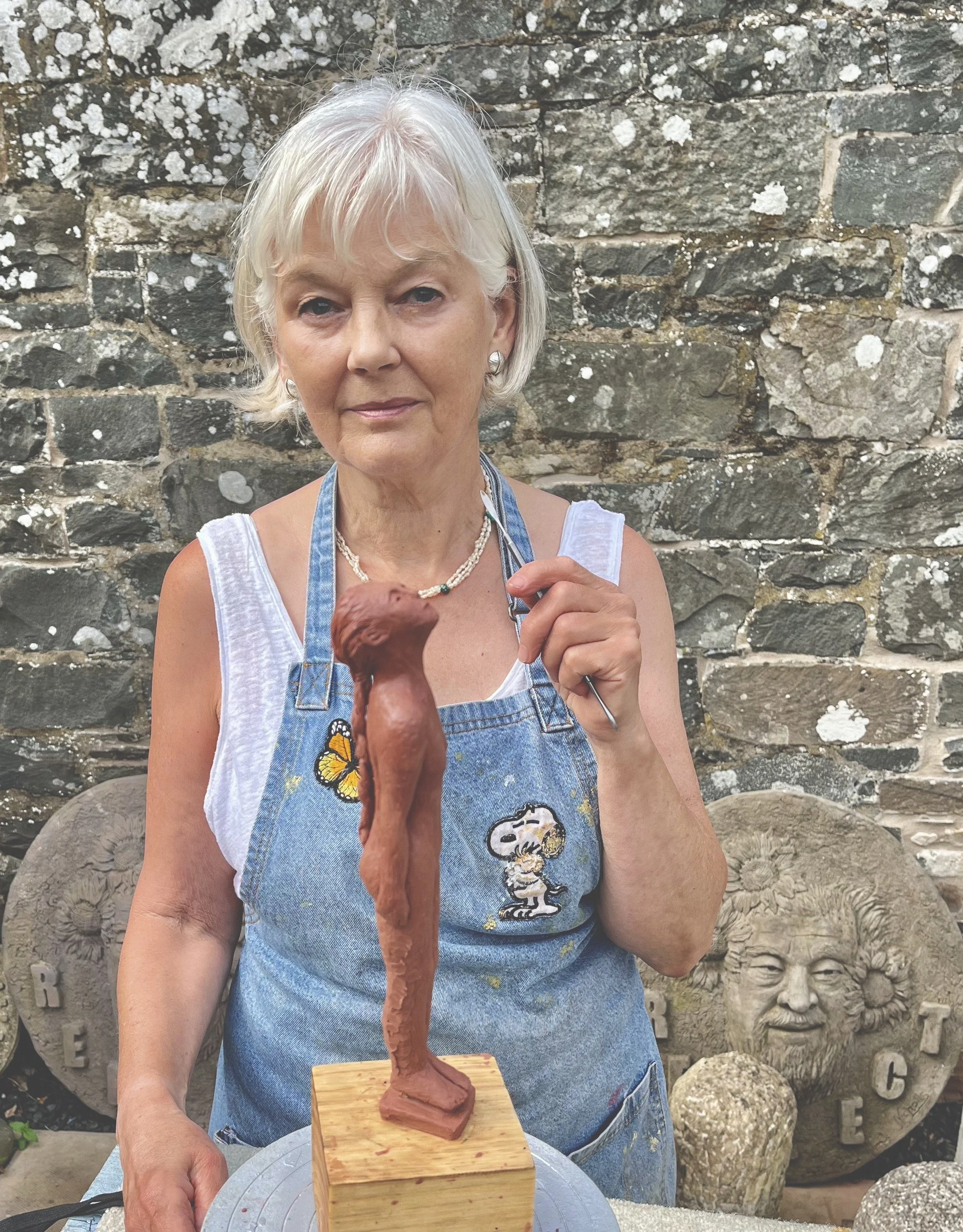 Photo of sculptor Deirdre Nicholls in her studio yard, holding a figure sculpted in wax, with sculptures in the background of Ai WeiWei in cast concrete.