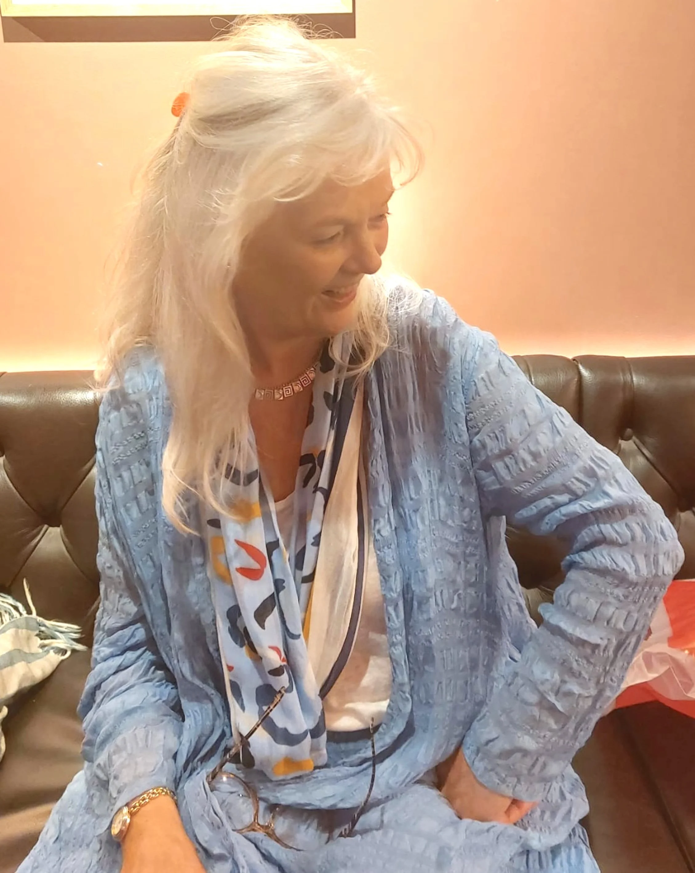 A woman with long, white hair smiling and looking down, sitting on a leather couch in a warmly lit room, wearing a light blue textured jacket, a patterned scarf, and a gold watch.