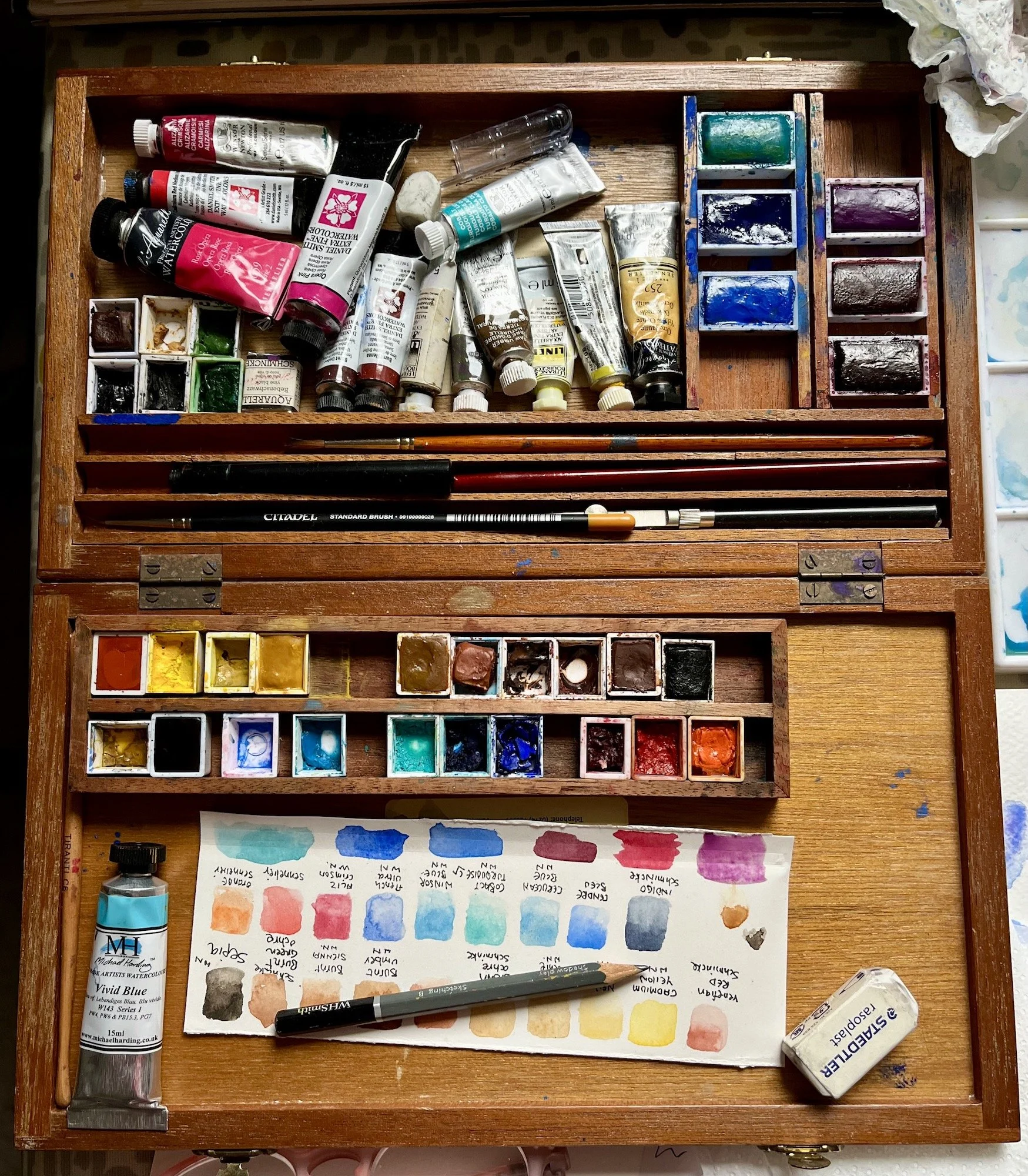 Vintage wooden paintbox containing watercolour paints in tubes & pans, together with brushes and pencils, on a background of Arches watercolour paper block
