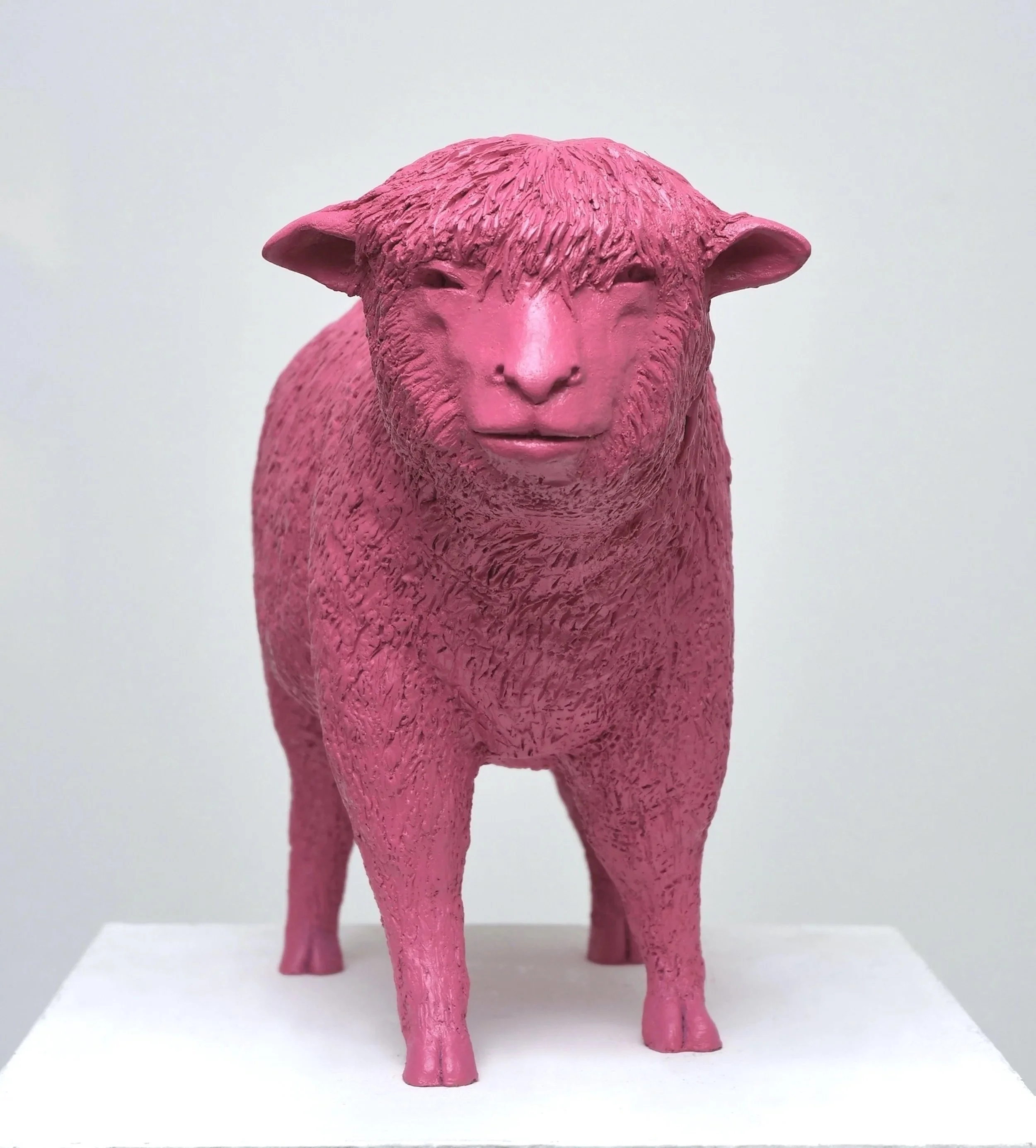 Pink sculpture of a sheep with a human-like face, standing on a white pedestal against a plain background.