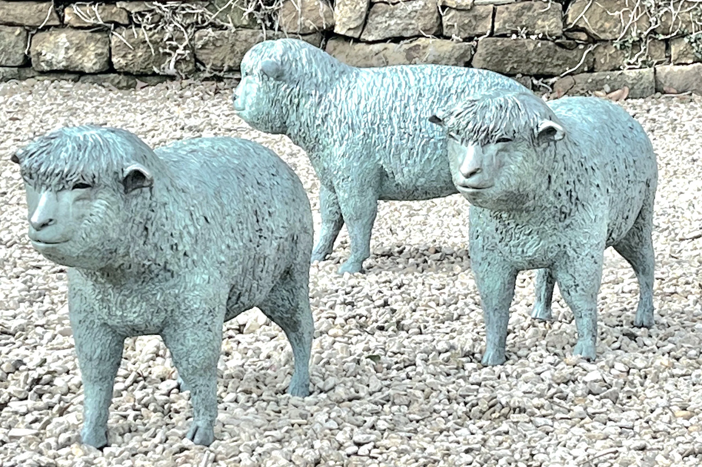 Three stone sheep statues on gravel ground with stone wall in background.