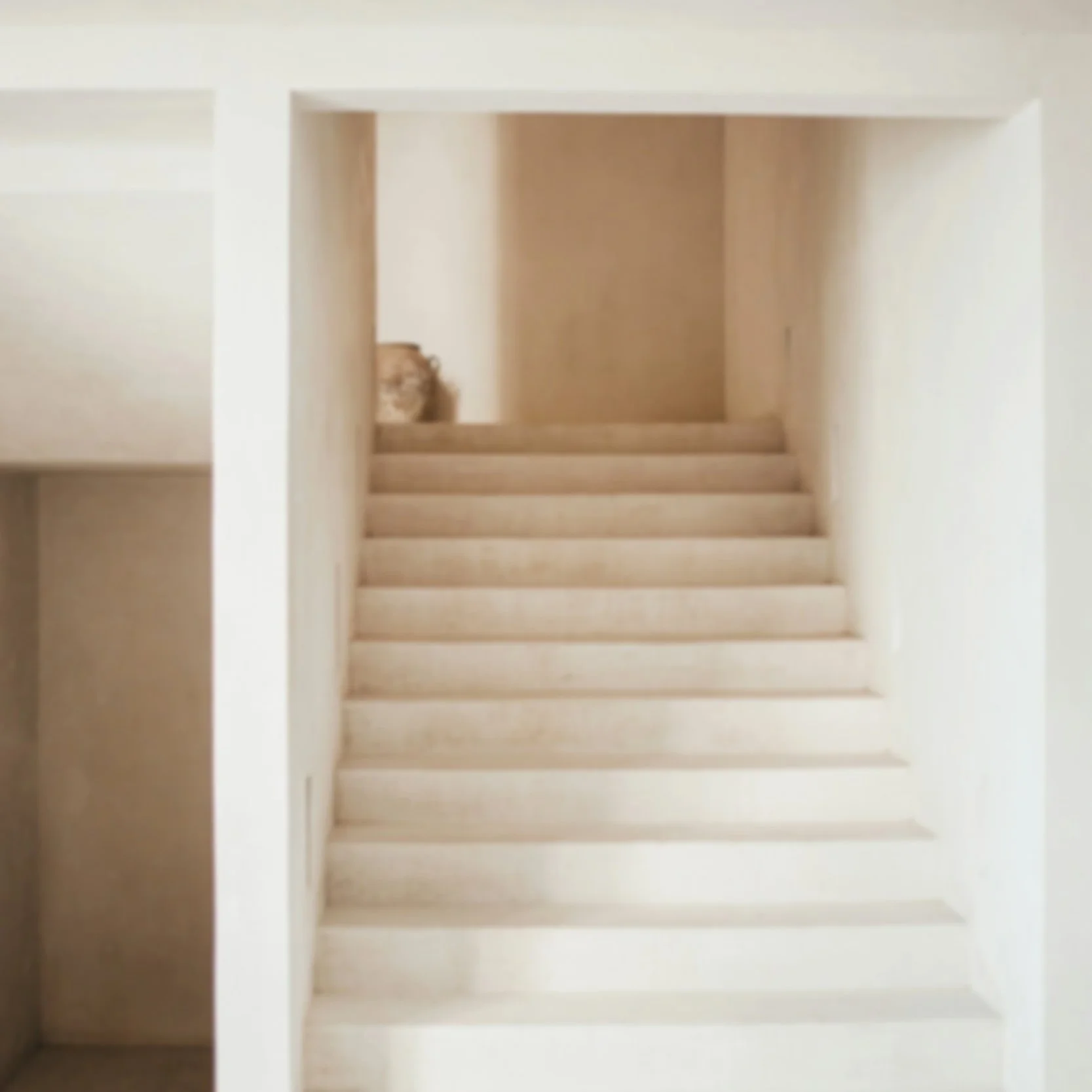 Innenansicht eines Gebäudes mit weißen Wänden und einer Treppe, die nach oben führt, mit einem kleinen braunen Objekt oder Skulptur auf der oberen Treppenseite.