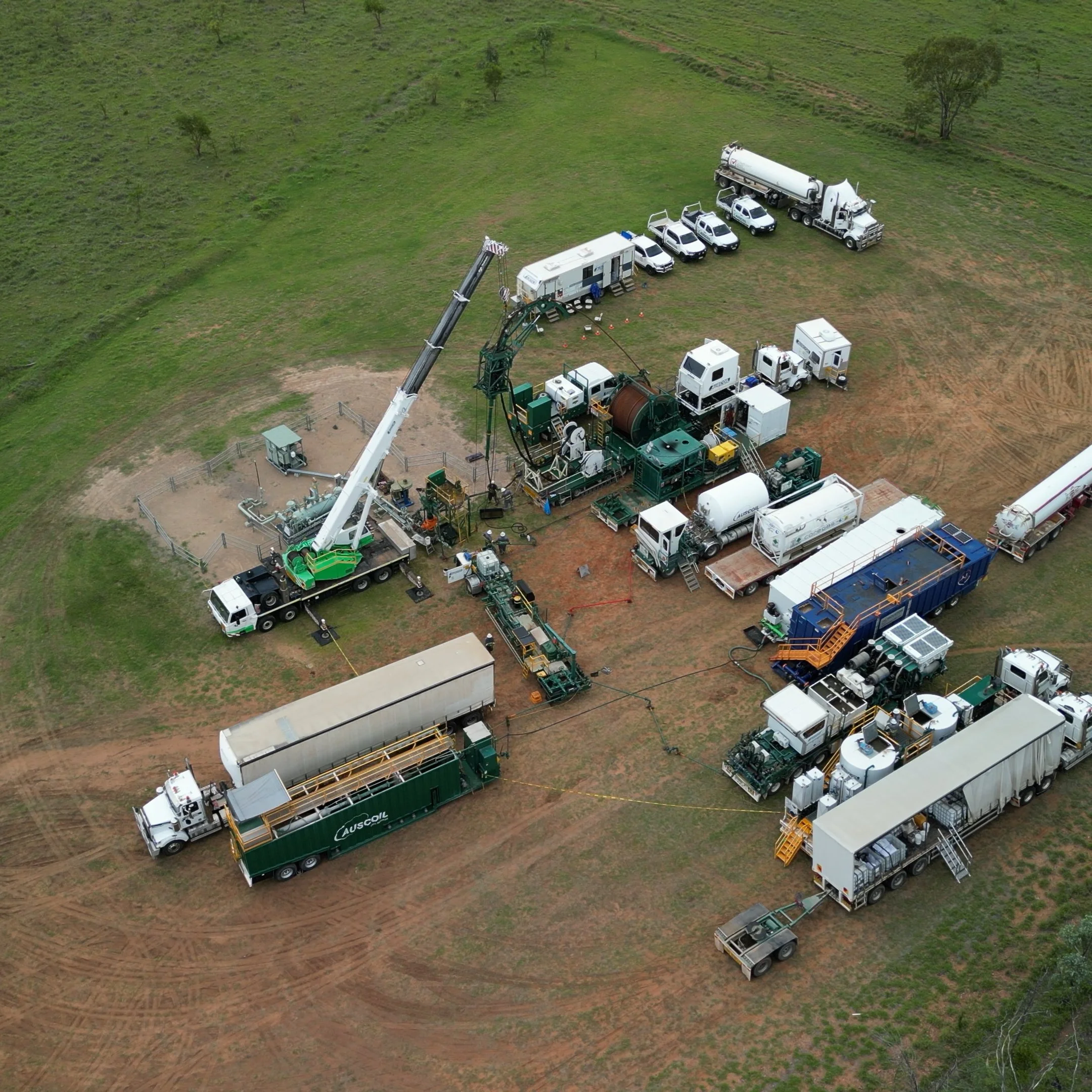 An aerial view of a construction or industrial site with multiple trucks, trailers, and equipment arranged in a circular pattern on a grassy area.