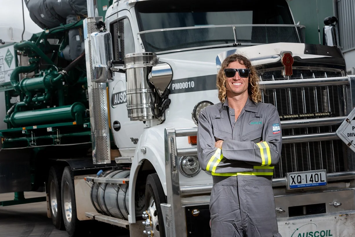 A person in a gray work jumpsuit and sunglasses is standing in front of a large white heavy-duty truck with green machinery on the side, smiling with arms crossed.