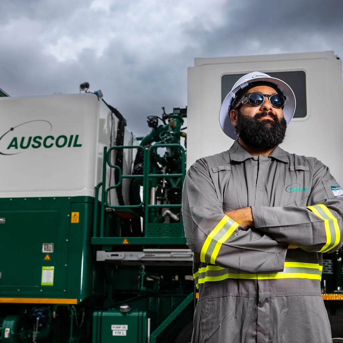 A man wearing sunglasses, a white safety helmet, and a gray uniform with yellow reflective stripes stands confidently with arms crossed in front of a large green industrial machine labeled 'AUSCOIL' under a cloudy sky.