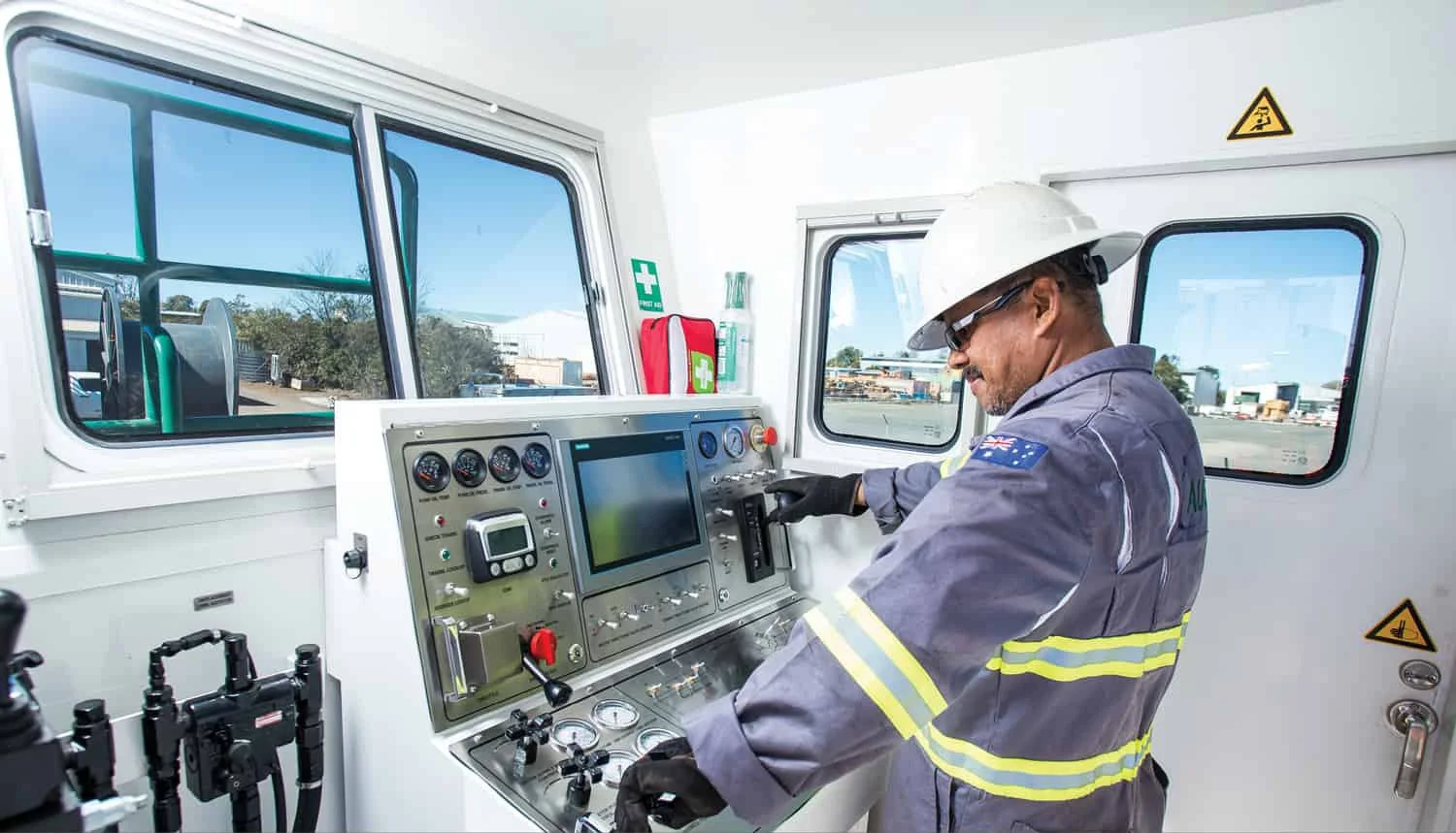 A man wearing safety glasses, a white hard hat, gloves, and a gray uniform operating controls inside an industrial control room with windows showing an outdoor industrial area.