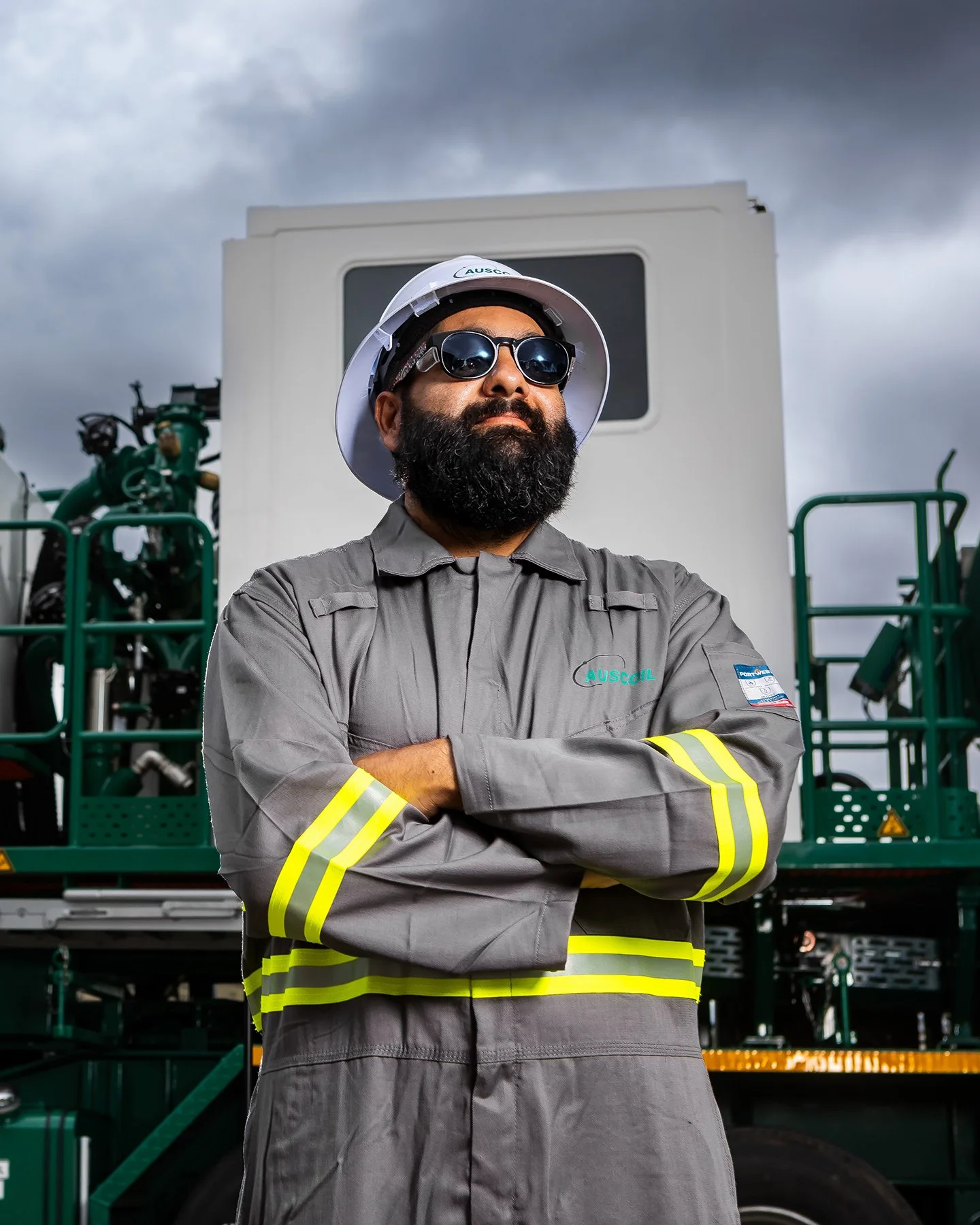 A man with a beard wearing sunglasses, a hard hat, and a gray safety uniform with reflective stripes standing in front of industrial machinery.