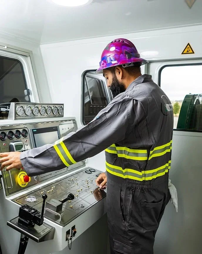 A worker operating controls inside a mobile control room or vehicle, wearing a purple safety helmet and gray coveralls with yellow reflective stripes.