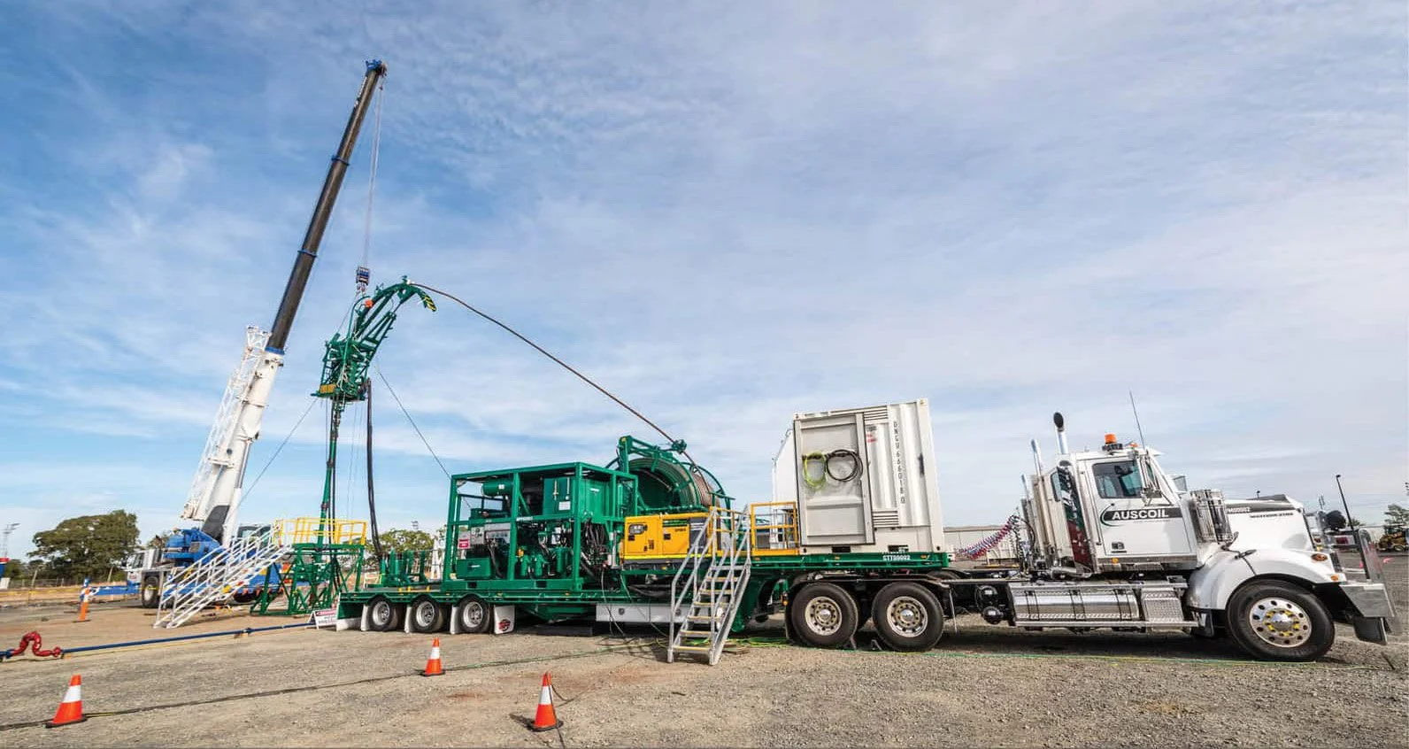 Large industrial drilling setup mounted on a white semi-truck with safety cones around the area, with a crane lifting equipment and a partly cloudy sky in the background.