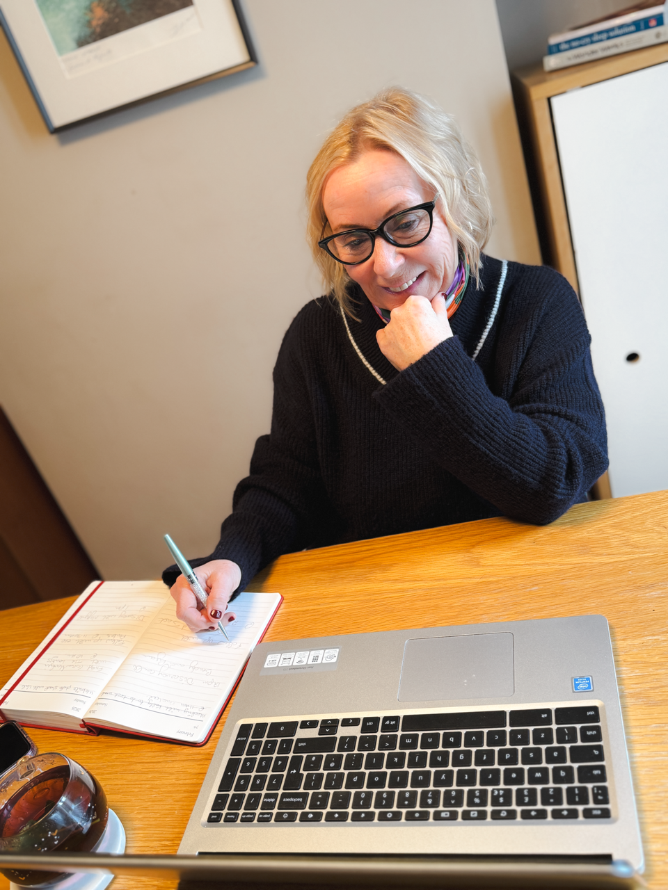 A woman with blonde hair, glasses, and a black sweater writing in a notebook at a wooden table, with a laptop and a glass of tea nearby.