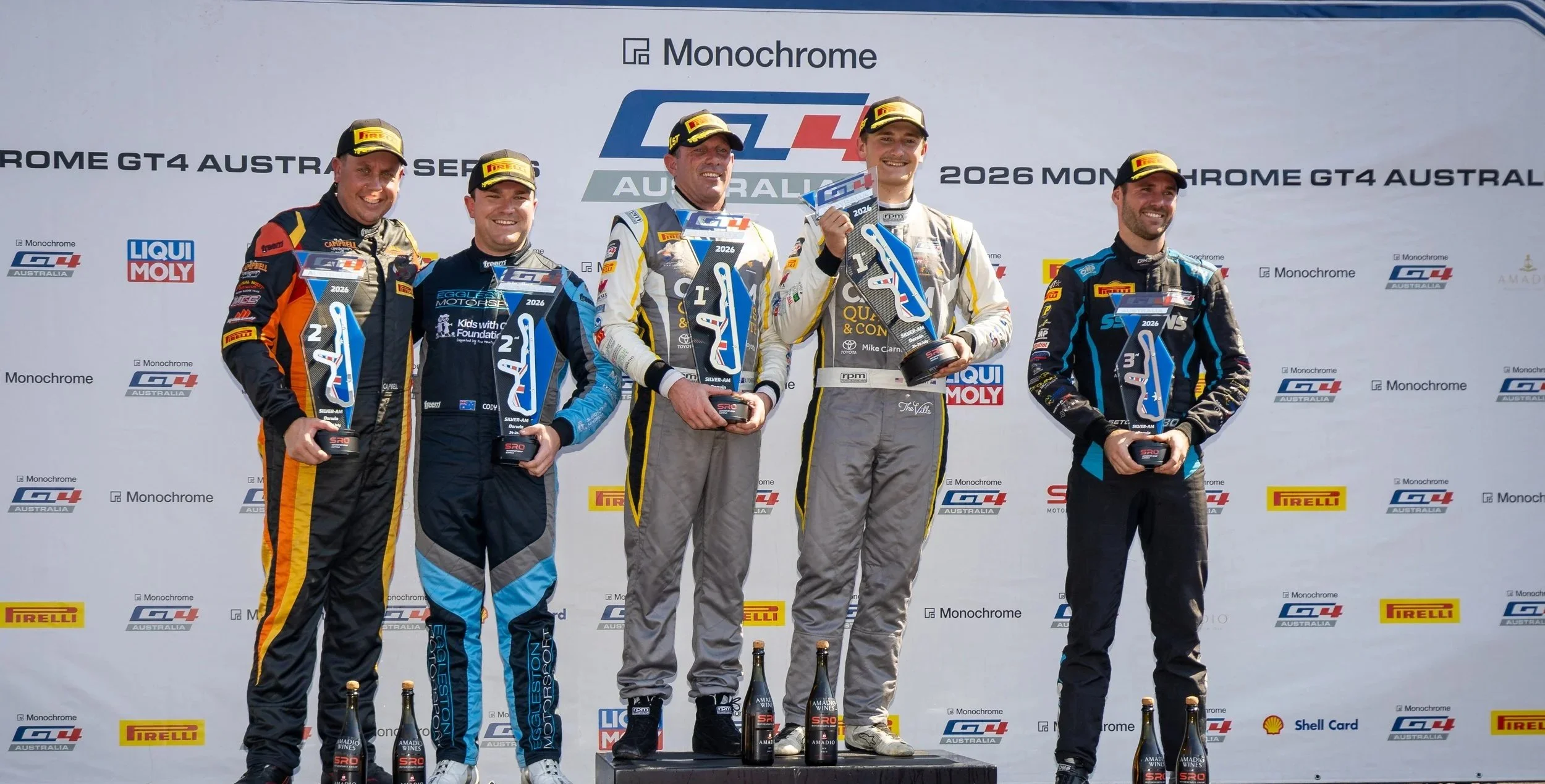 Race car drivers standing on the winners' podium celebrating their victory at the 2026 Monochrome GT4 Australia Series, holding trophies and champagne bottles.