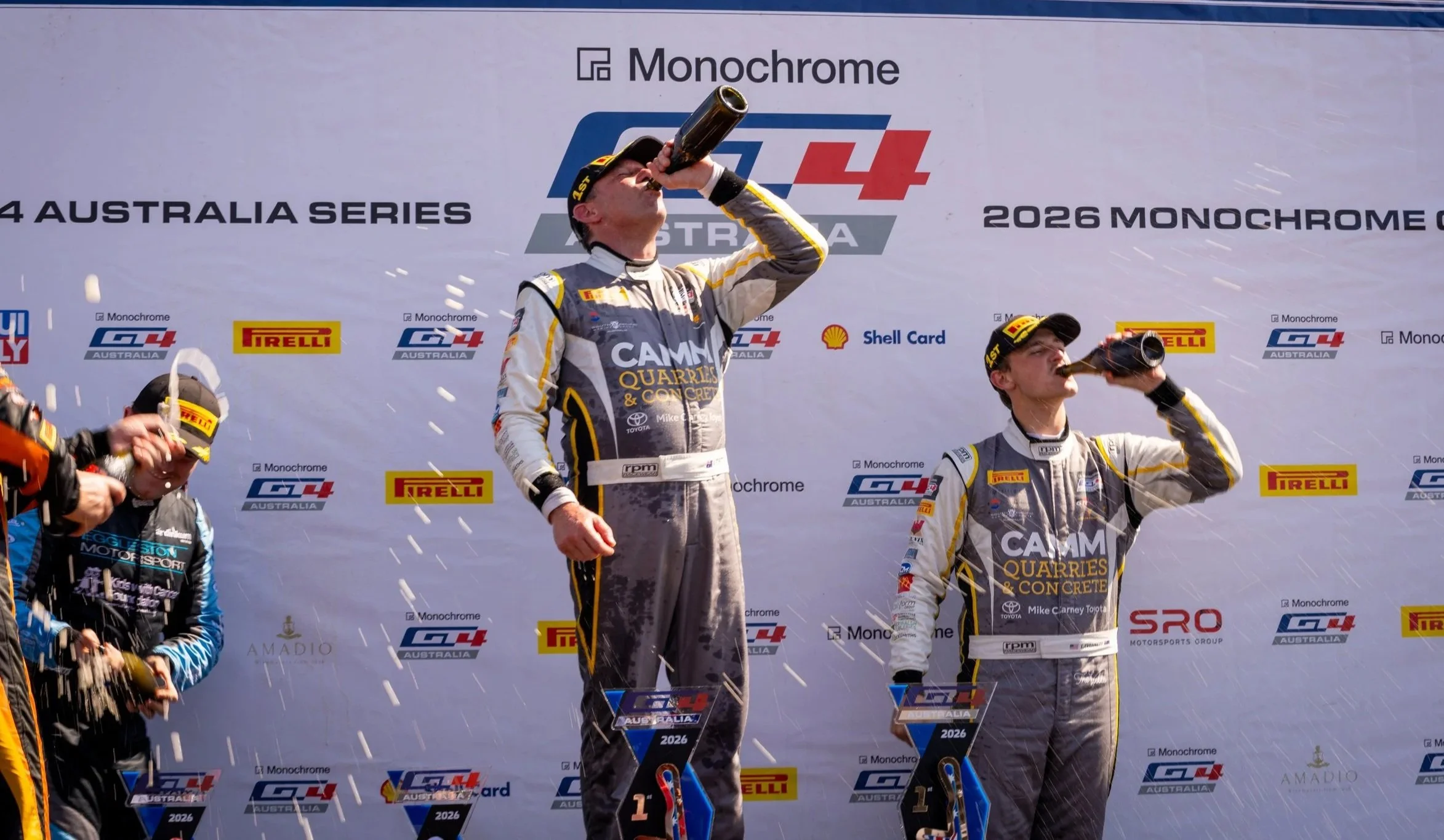 Race car drivers on a podium celebrating with champagne at the 2026 Monochrome GT4 Australia Series, with one driver drinking from a bottle, standing in front of a branded backdrop with event and sponsors logos.