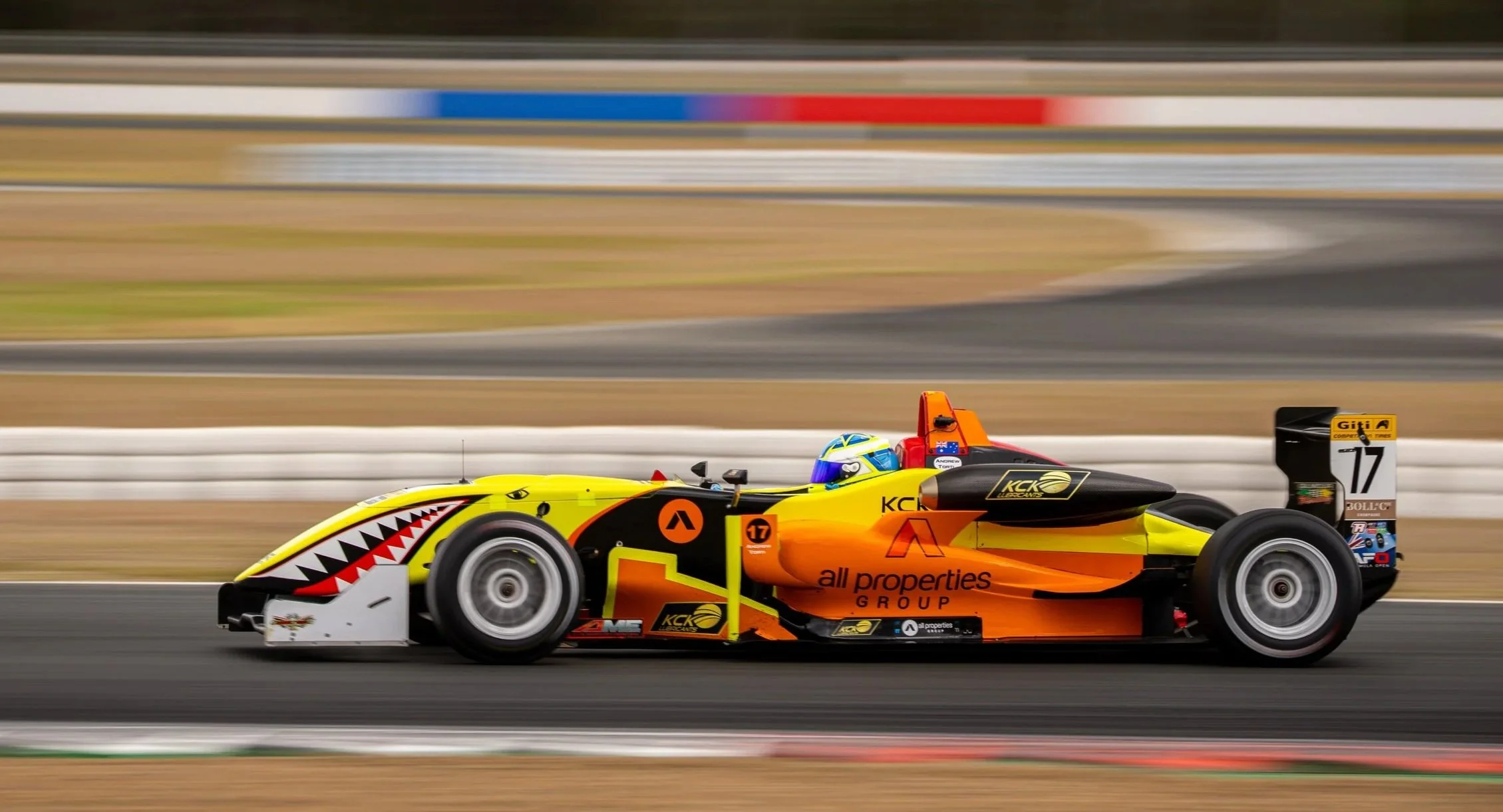 A yellow, black, and orange open-wheel race car on a racetrack, with a shark mouth design on the front and various sponsor logos, driven by a driver wearing a blue helmet.